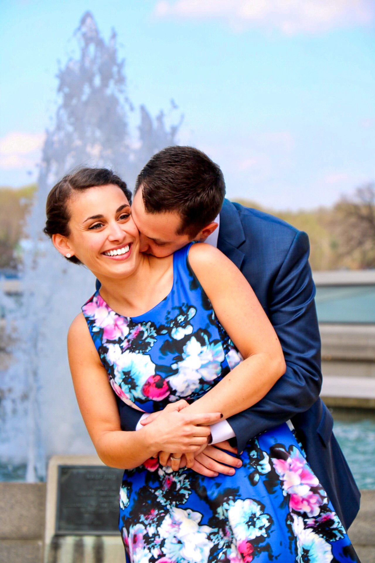 A smiling woman in a blue floral dress is being embraced from behind by a man in a blue suit, who is kissing her on the neck near a fountain.