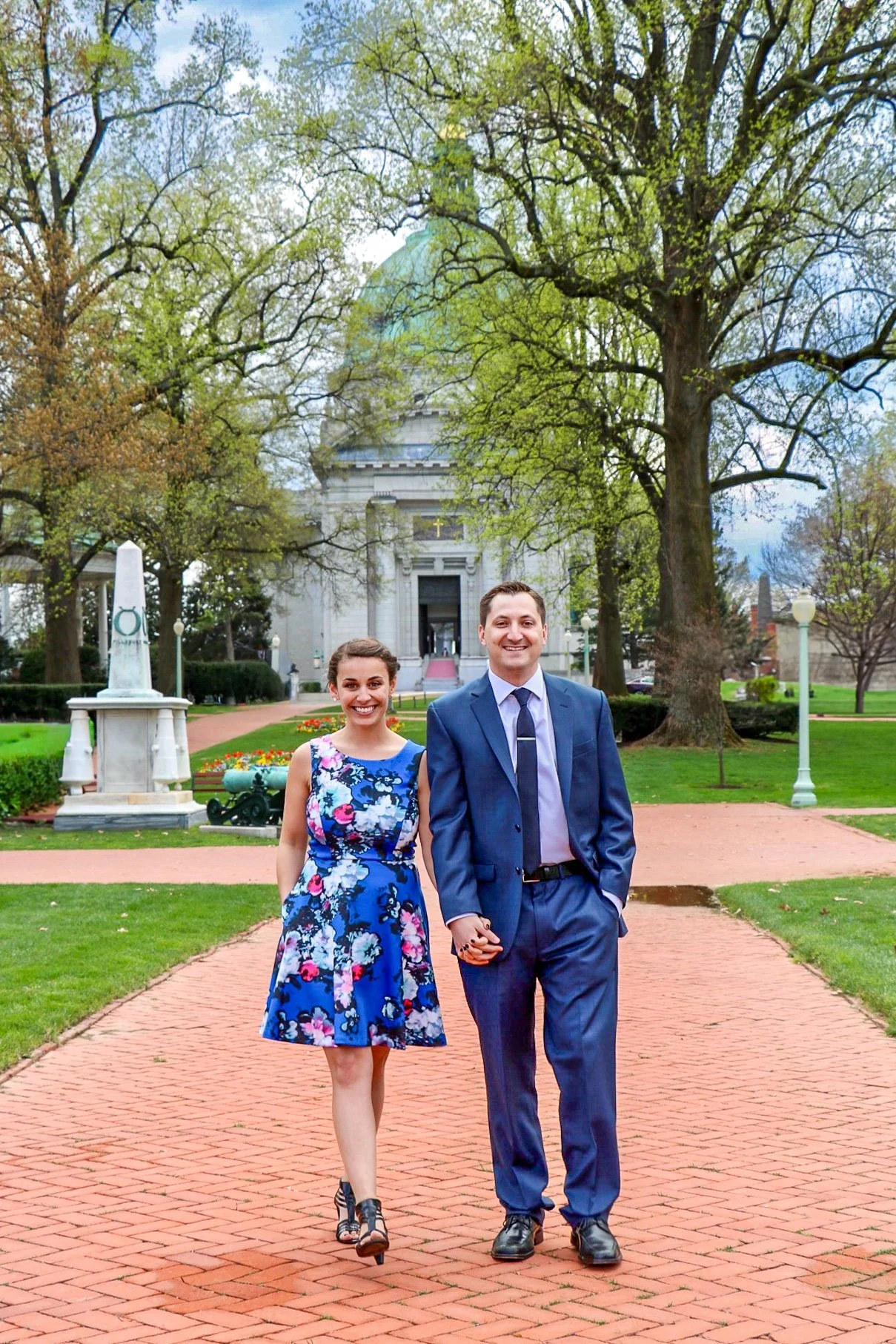 A young woman and man in formal attire walking hand-in-hand on a brick path in a park with green grass, trees, lamp posts, and a large historic building in the background.
