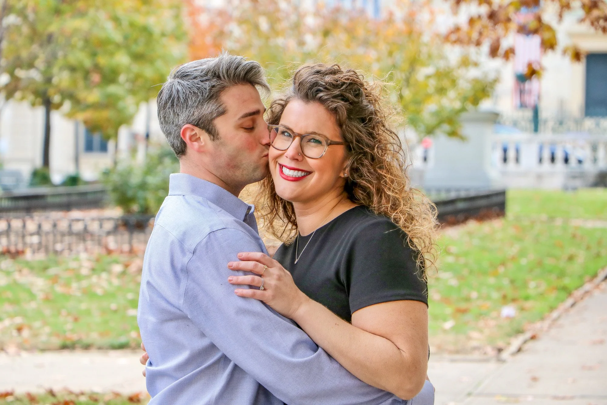 A man kissing a woman on the cheek outdoors during autumn, with colorful fall foliage in the background.