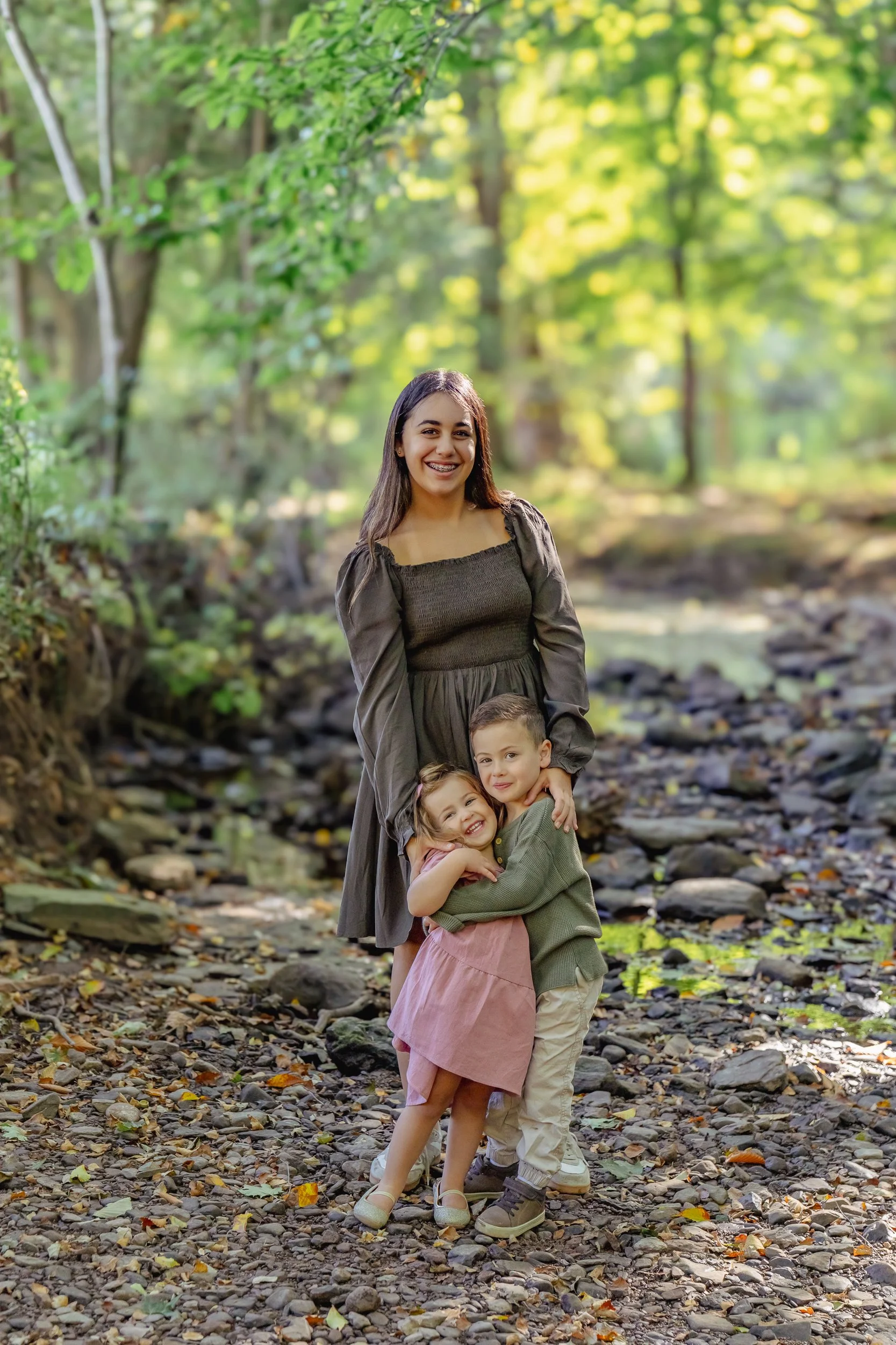 A teenager and two young children standing close together on a rocky path in a forest with green foliage. The woman is smiling and wearing a brown dress, while the children are hugging her, smiling happily.