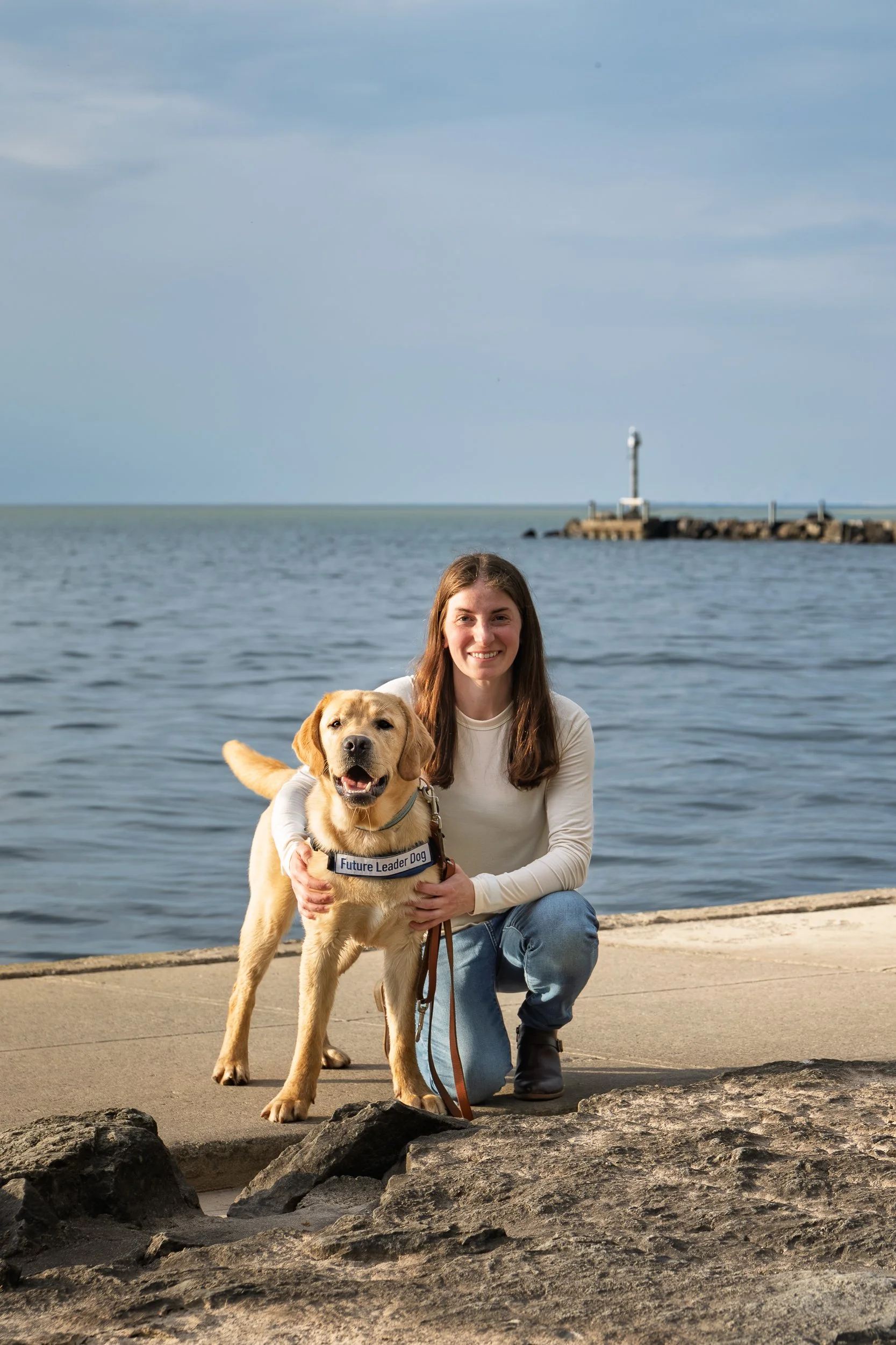 Puppy raiser and yellow Labrador posing on the rocky shoreline at Webster Park with Lake Ontario and a lighthouse in the background, Webster NY