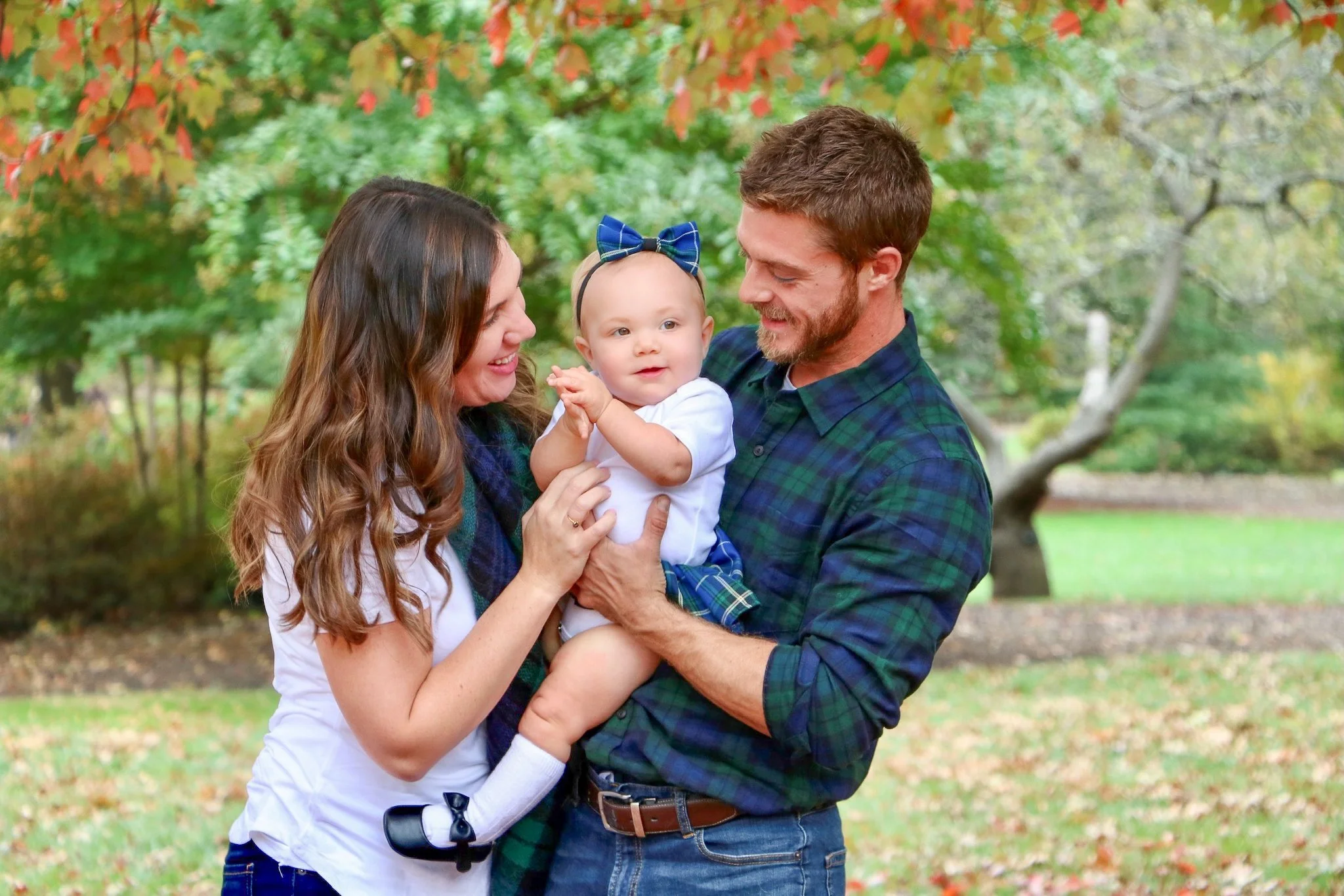A happy family of three, a woman, a man, and a baby girl, standing outdoors in a park with autumn foliage. The woman and man are smiling at the baby, who is wearing a white shirt, plaid shorts, and a blue bow on her head.