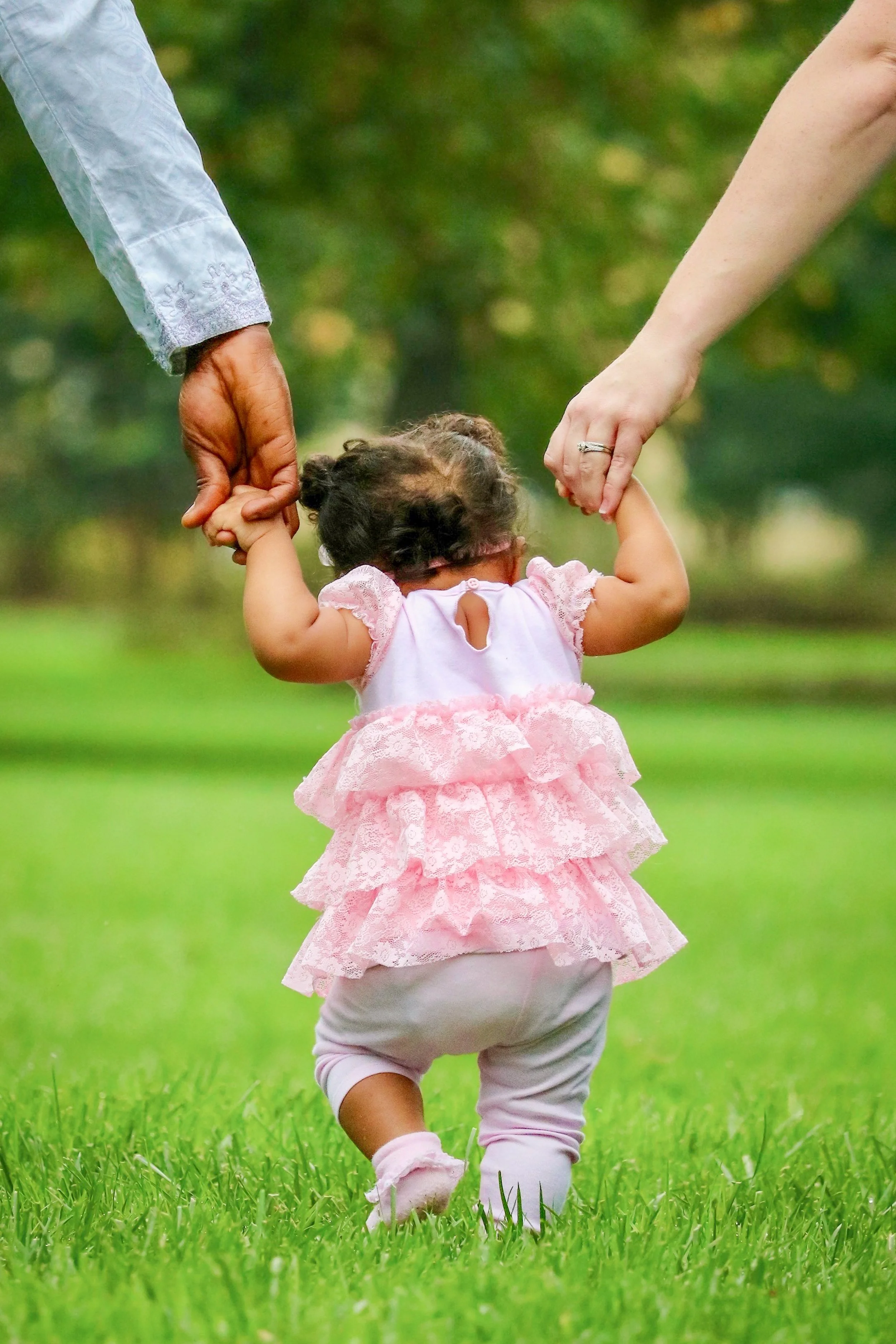 A toddler girl dressed in pink, holding hands with her parents as she takes steps on grass in a park.