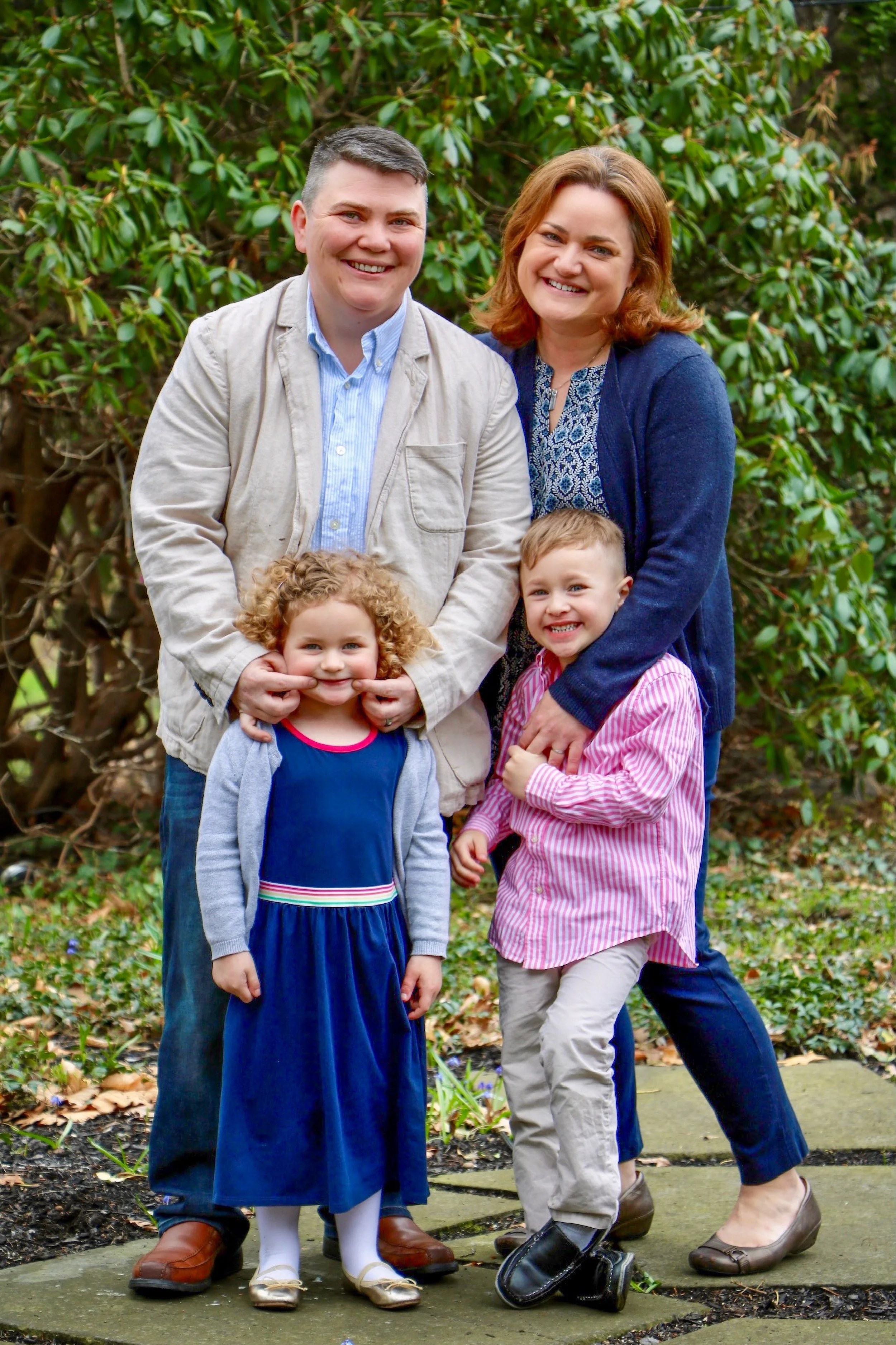 A family of four posing outdoors on a stone path surrounded by greenery, including a two women, a girl, and a boy, smiling at the camera.