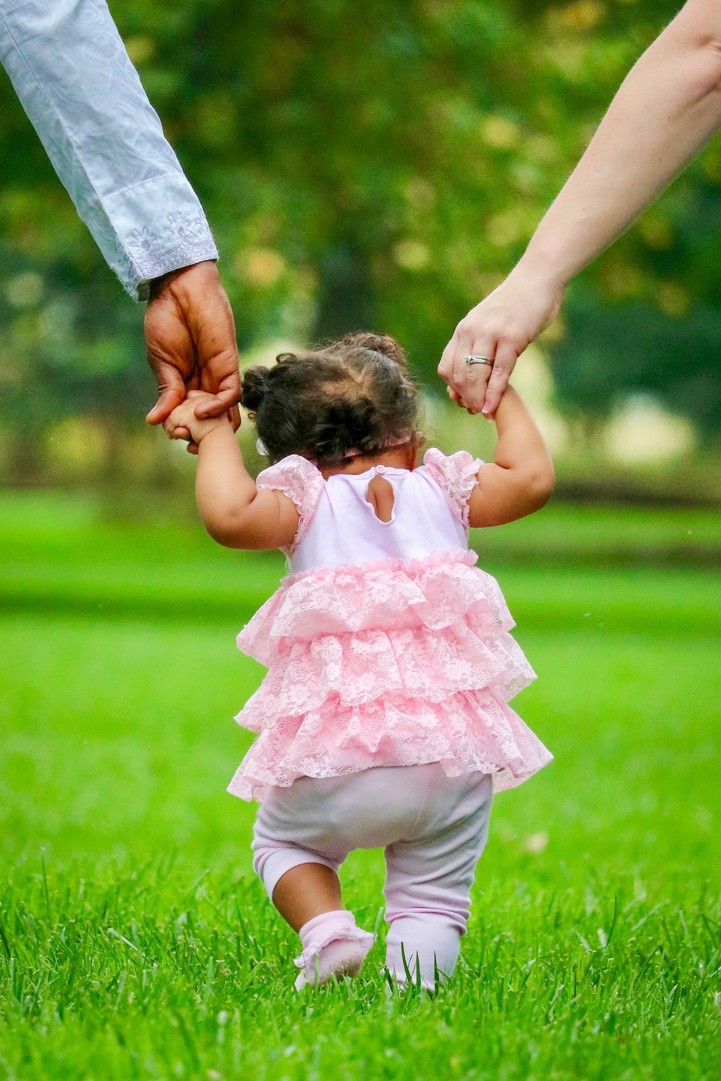 A toddler girl dressed in pink, holding hands with two adults as she takes steps on grass in a park.