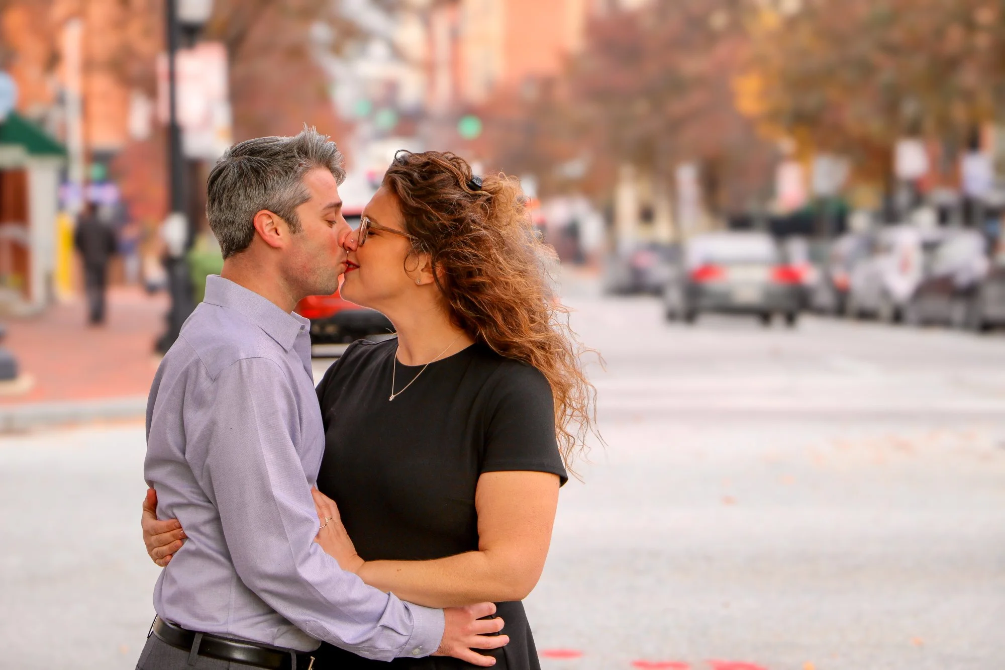 A couple is sharing a kiss on a city street in fall, with trees and parked cars in the background.
