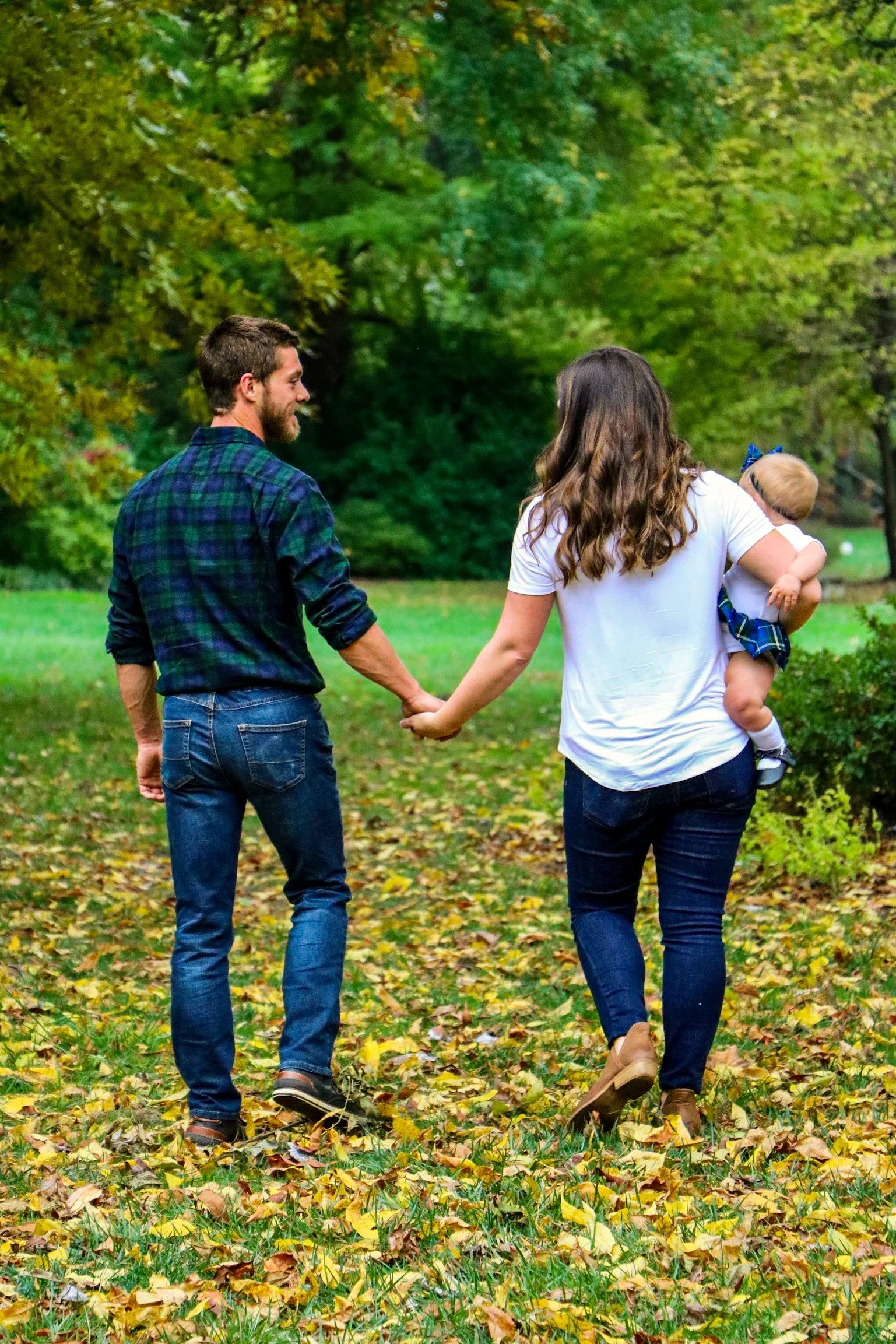 A family walking hand in hand through a park with green trees and fallen leaves, with a woman holding a young child.
