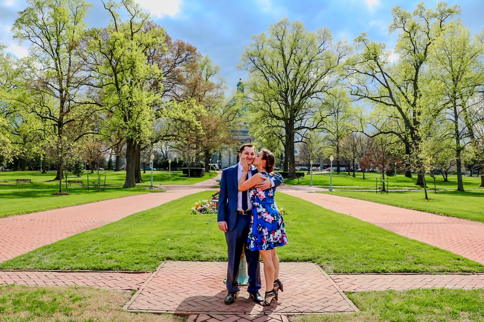 A couple dressed in formal clothes sharing an affectionate moment in a park with green grass and tall trees, with a building and cloudy sky in the background.