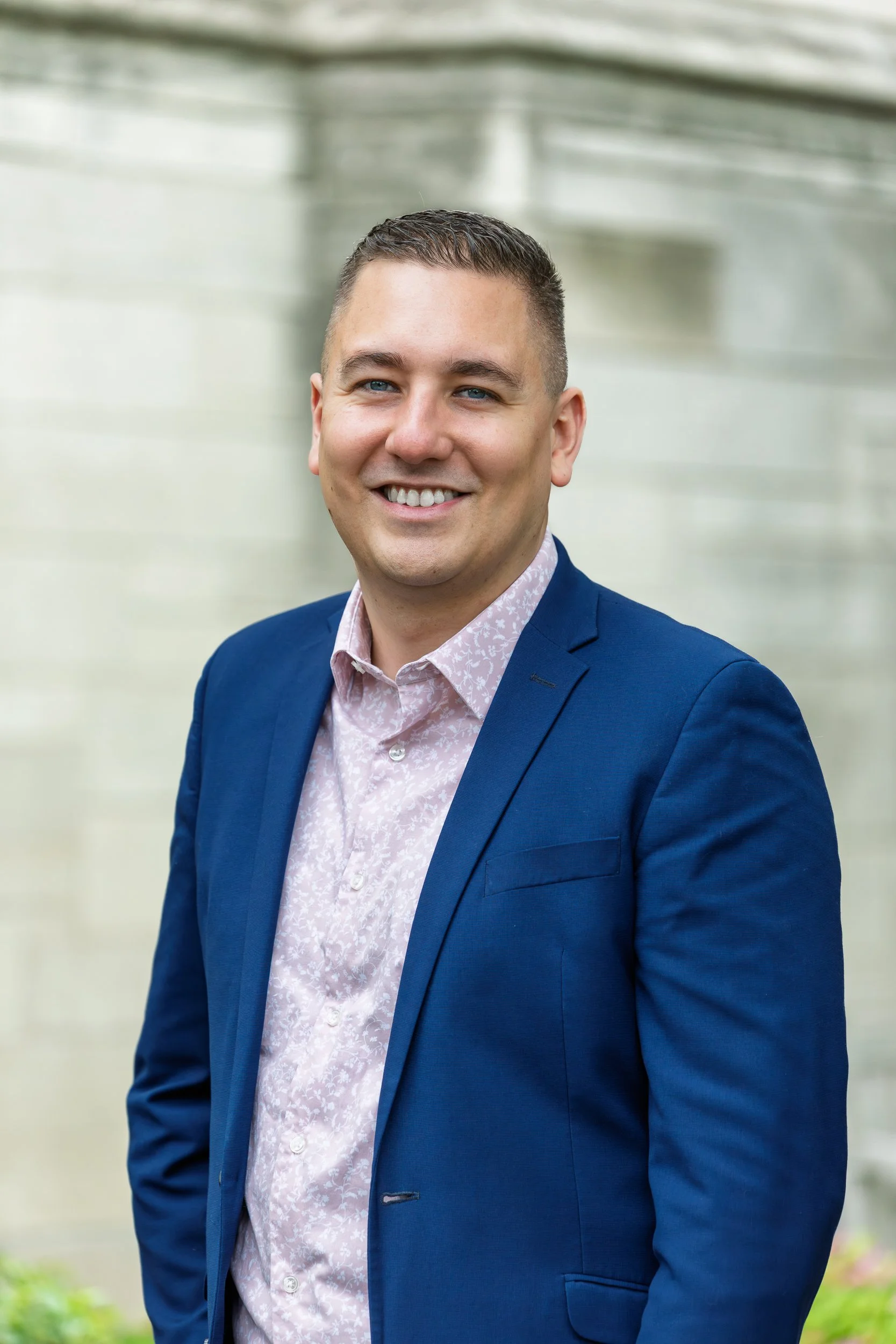 Professional headshot of a man in a navy blazer and floral dress shirt, smiling outdoors in front of a stone building