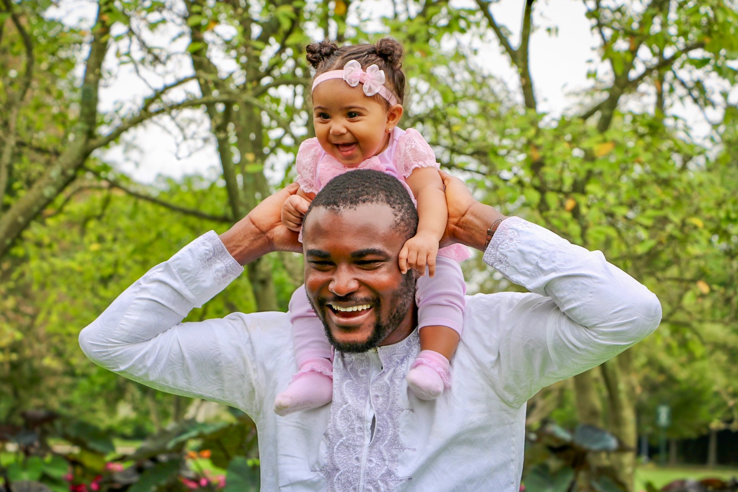 A joyful man carrying a young girl on his shoulders in a park, with green trees and foliage in the background.