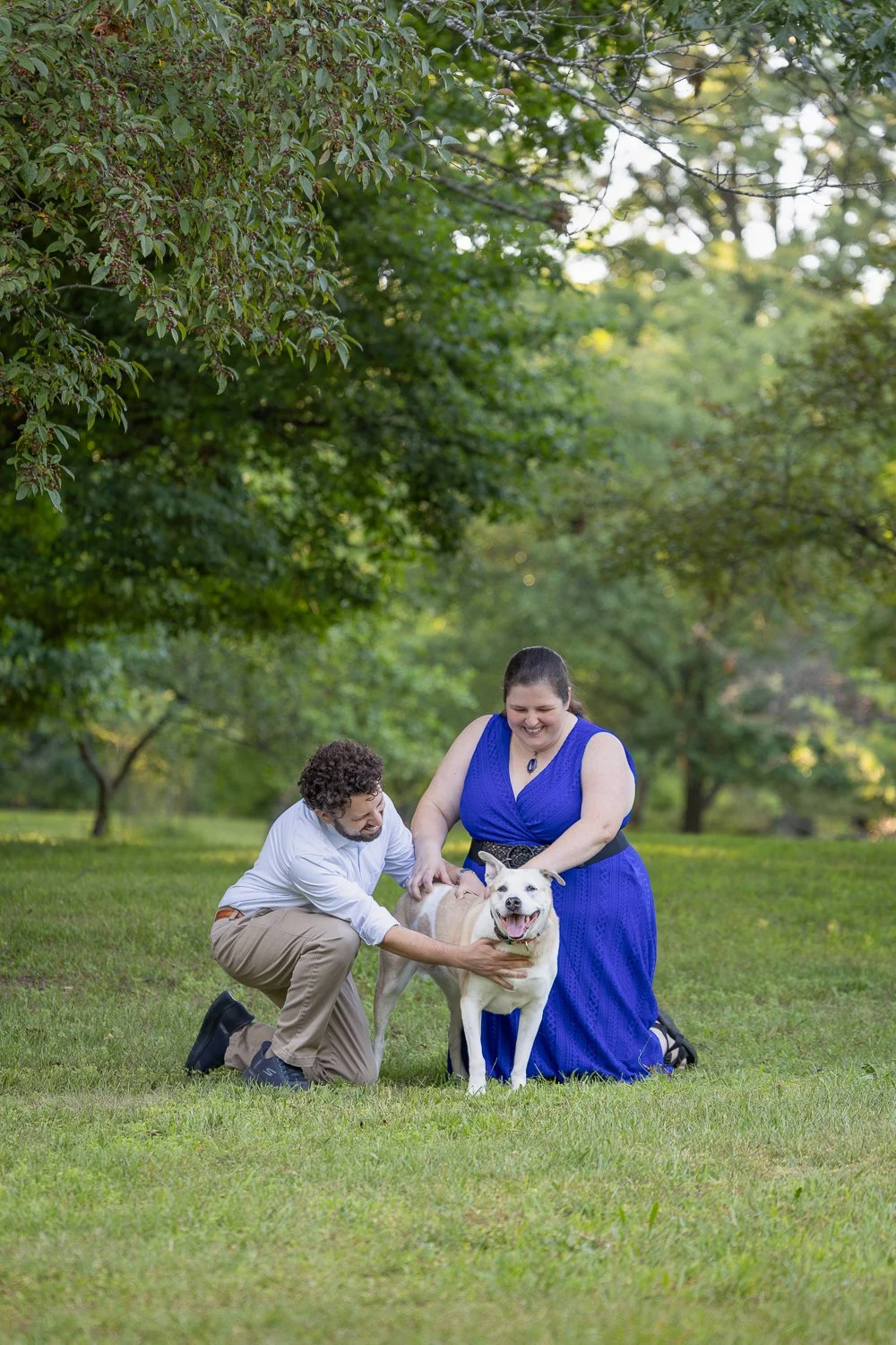 Couple kneeling on the grass at Durand Eastman Park in Rochester, NY, petting their dog together during a family pet photography session, surrounded by lush green trees.