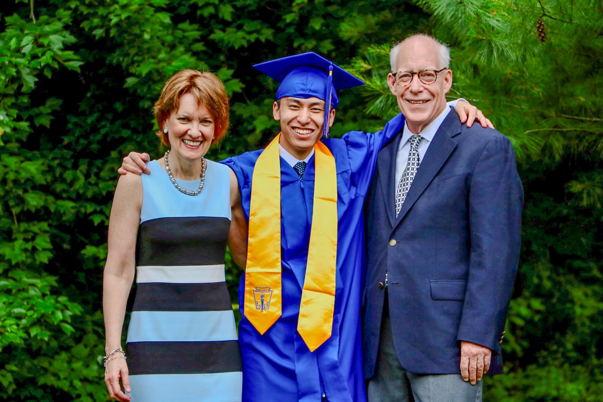 A young man in a blue graduation cap and gown, with a yellow stole, stands with his parents, a smiling woman in a striped dress and a man in a suit, all outdoors with green foliage in the background.