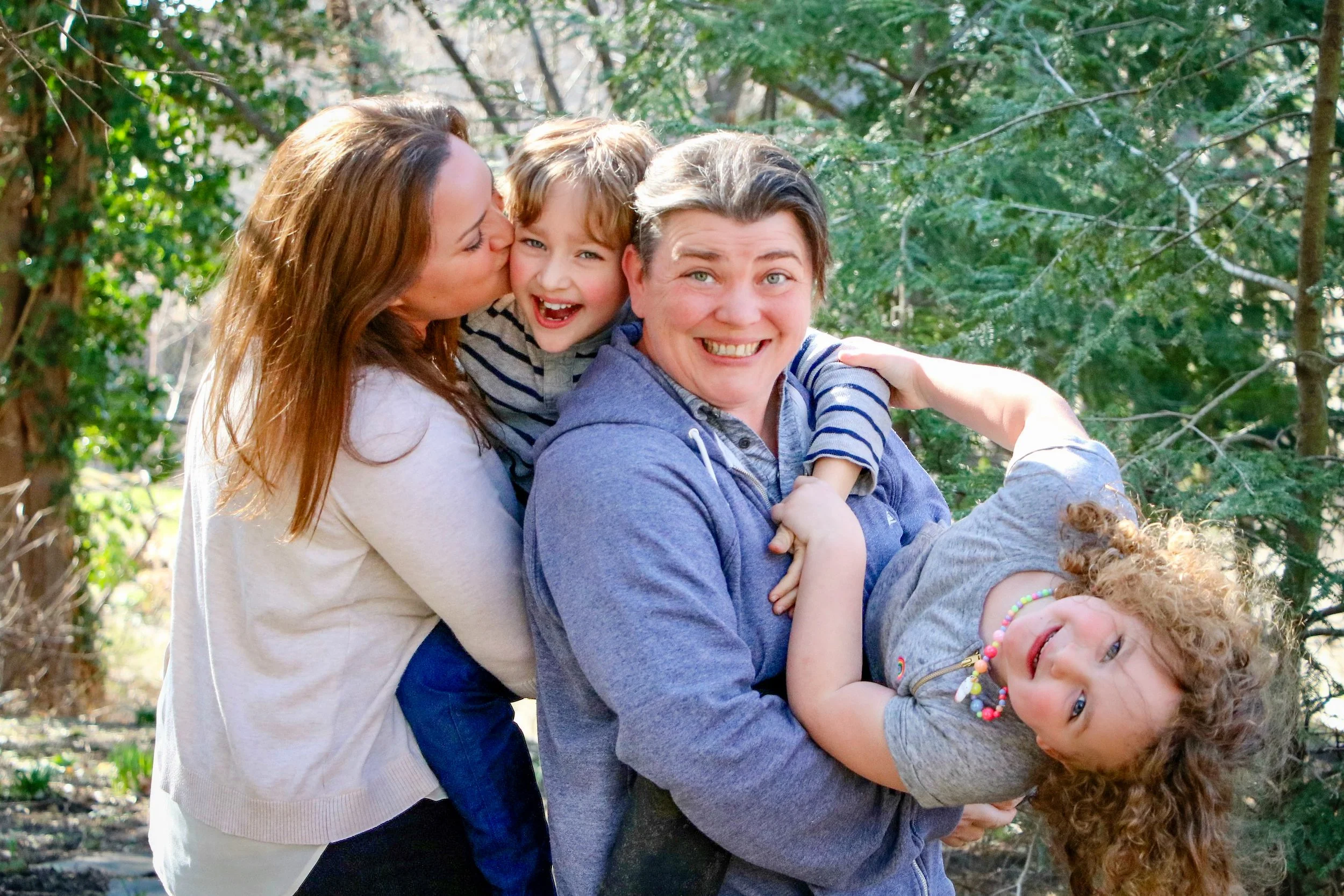 An active family portrait showing one mother kissing a young boy on the cheek and the other mother holding a playful young girl, surrounded by trees.