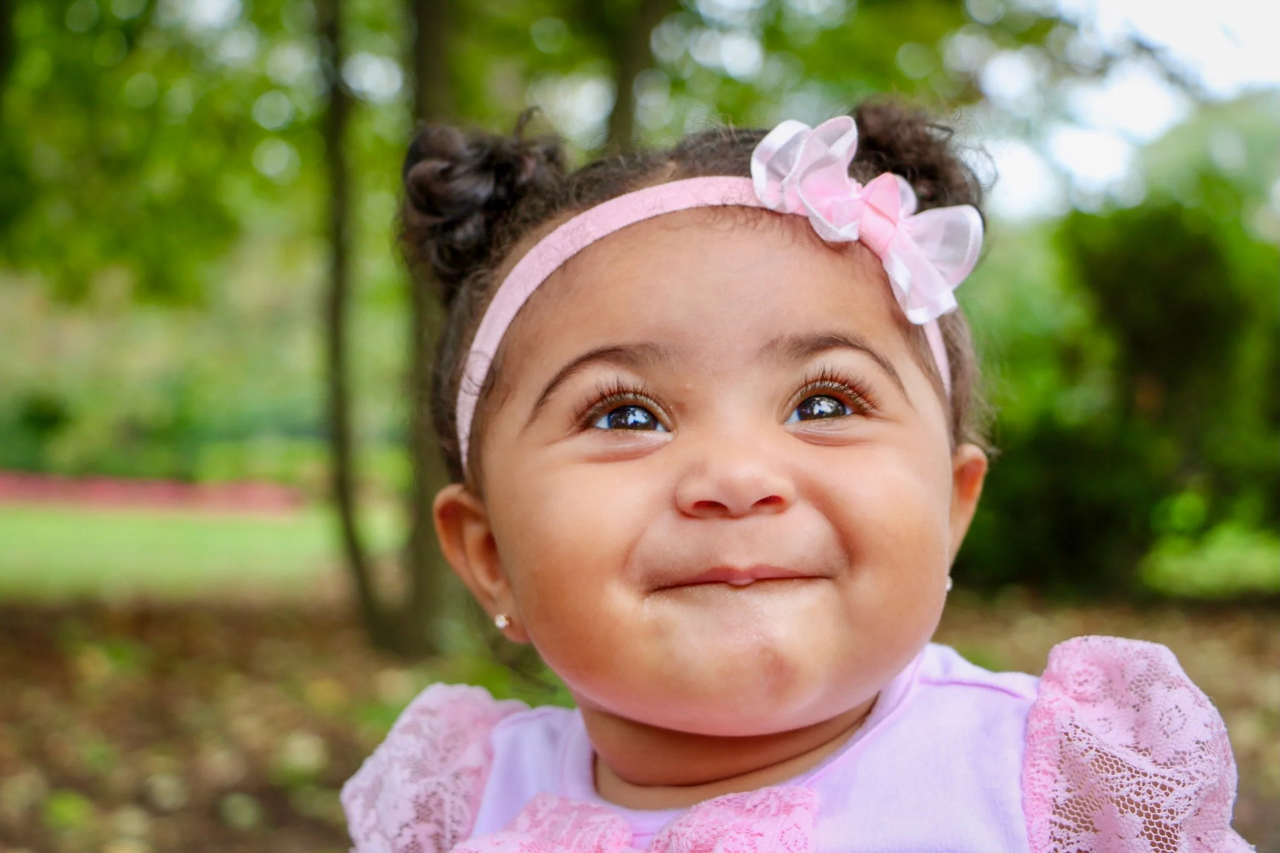 Close-up of a young girl with brown skin, dark curly hair in two buns, wearing a pink headband with a bow, smiling outdoors in a park with green trees in the background.
