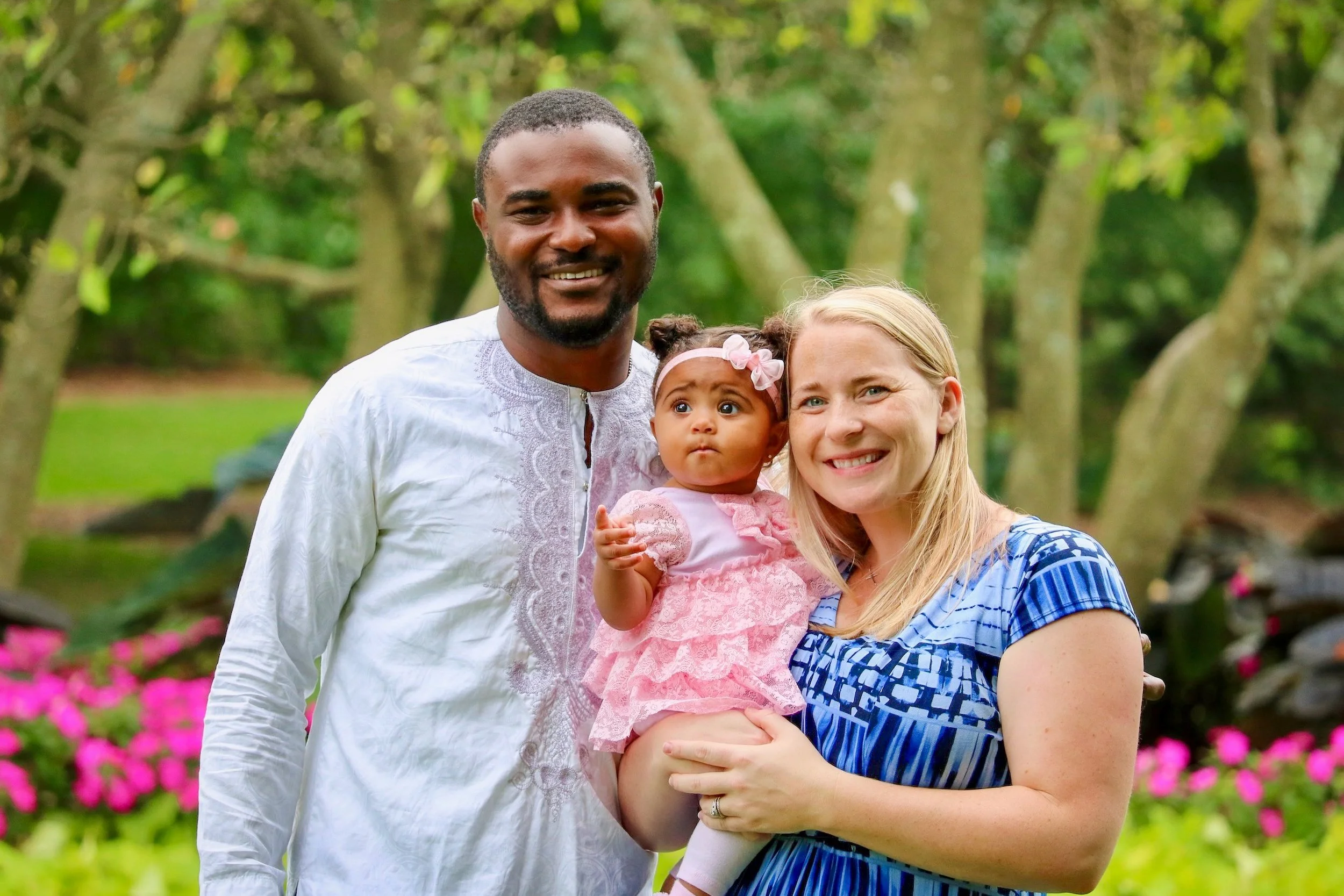 A diverse family of three, a man, a woman, and a young girl, smiling outdoors in a lush garden with green trees and pink flowers in the background.