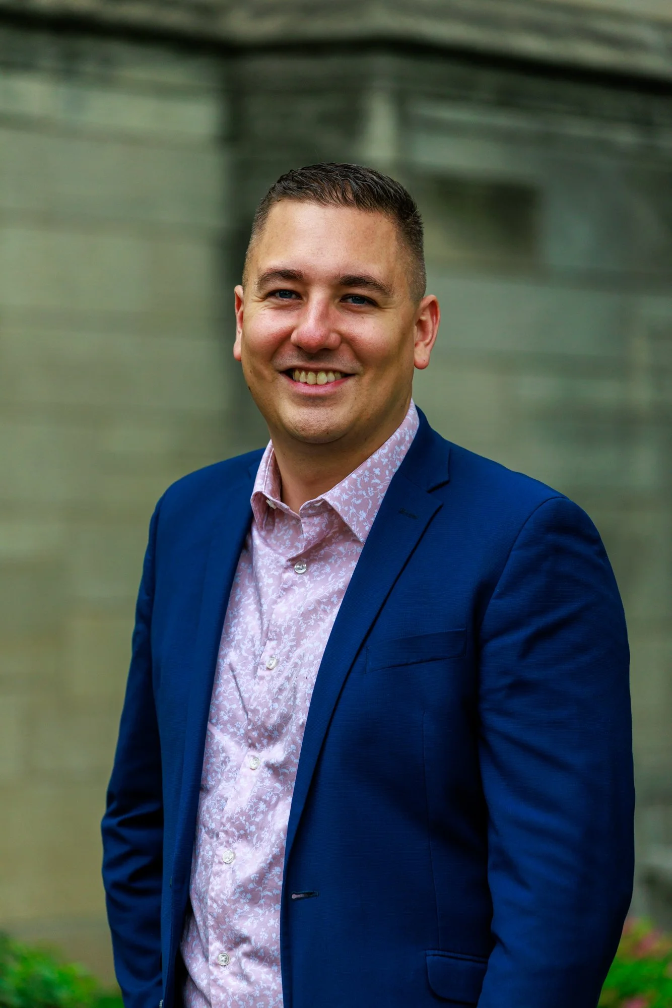 A smiling man in a blue suit and pink patterned shirt standing outdoors with a blurred building in the background.
