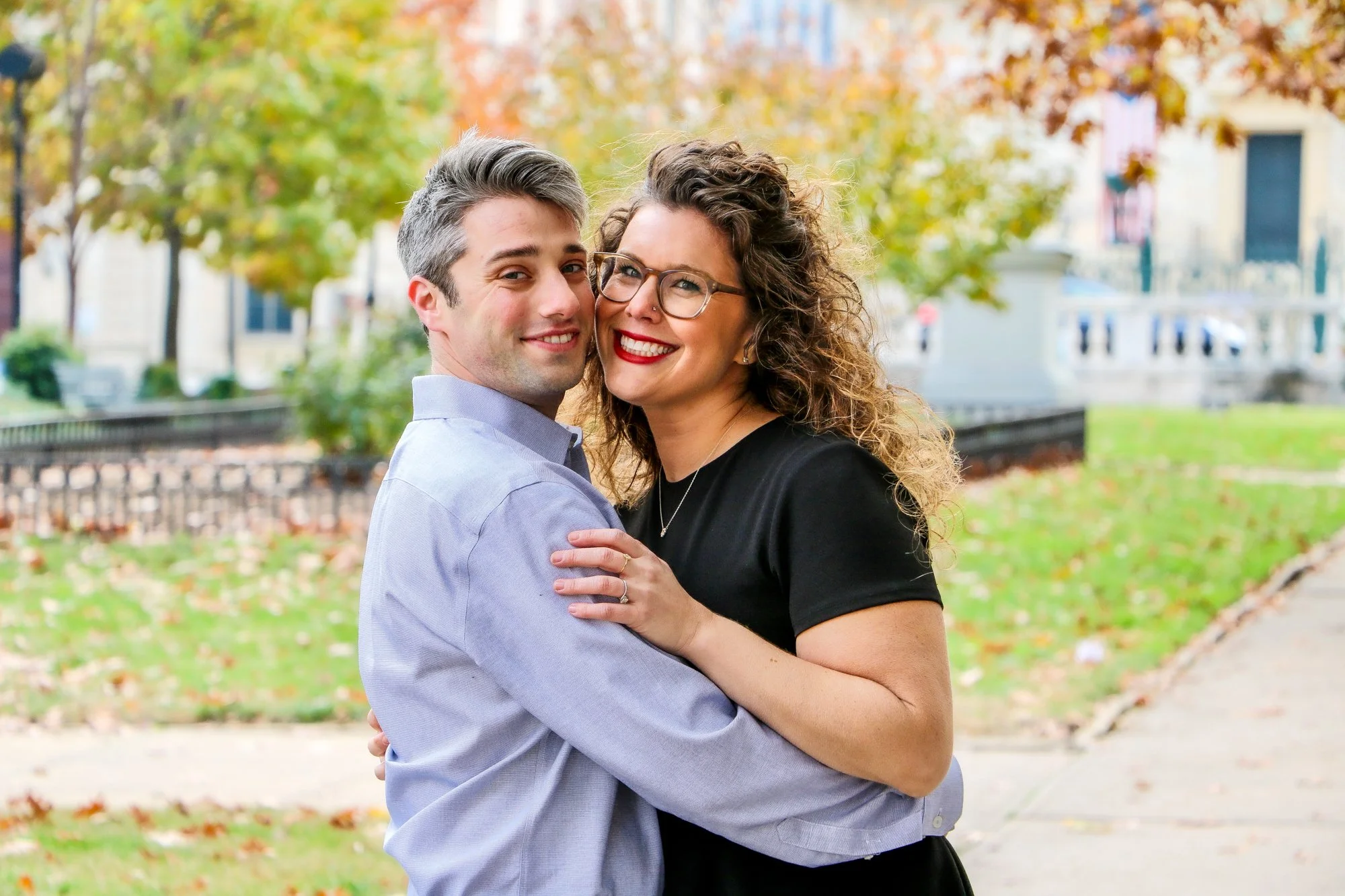 A happy couple hugging outdoors on a fall day, with trees and a sidewalk in the background.