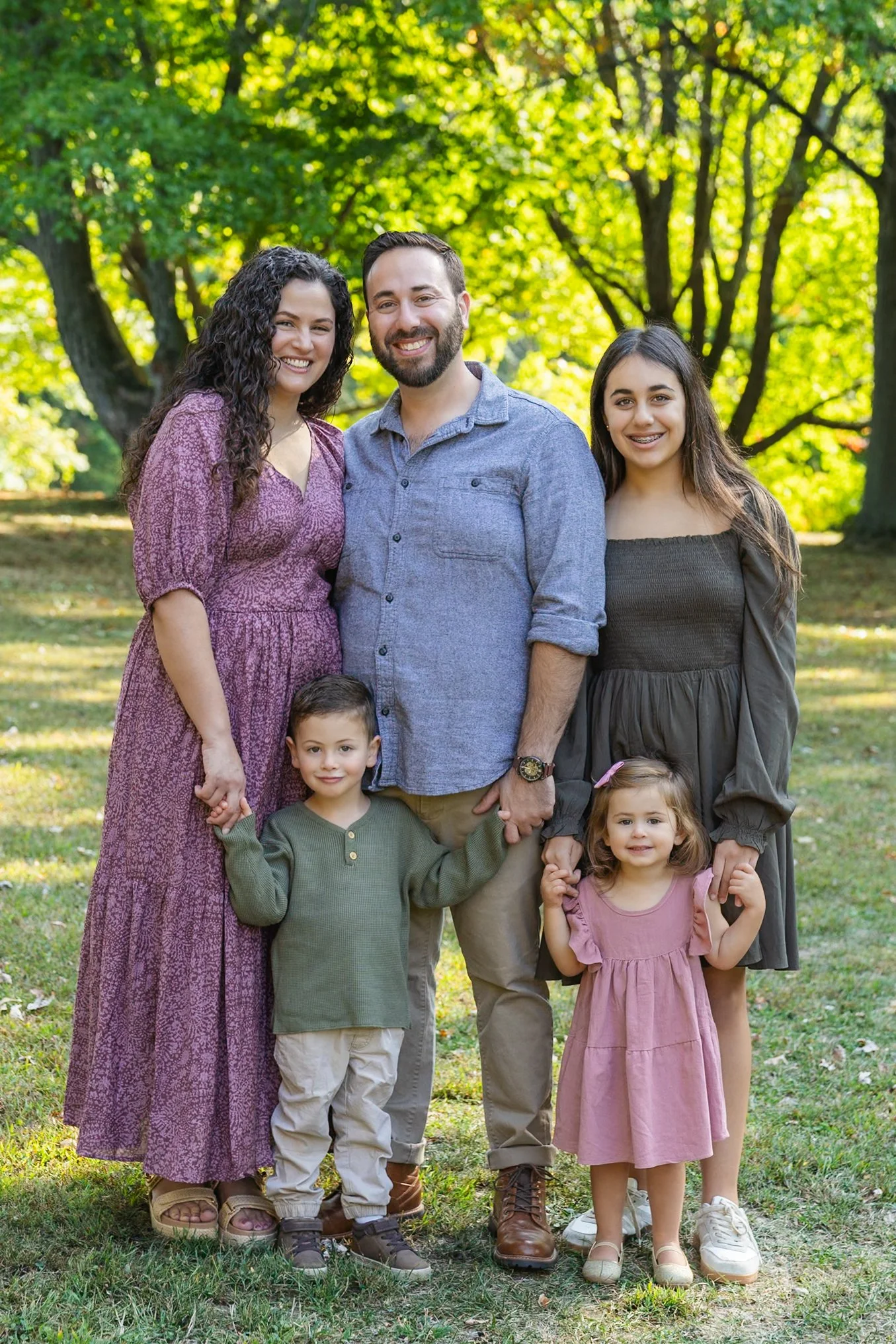 Family of five sitting outdoors in a park during fall, smiling at the camera. The family includes a man with a beard, a woman with curly hair, a teenage girl, a young boy, and a baby girl, with trees and autumn leaves in the background.