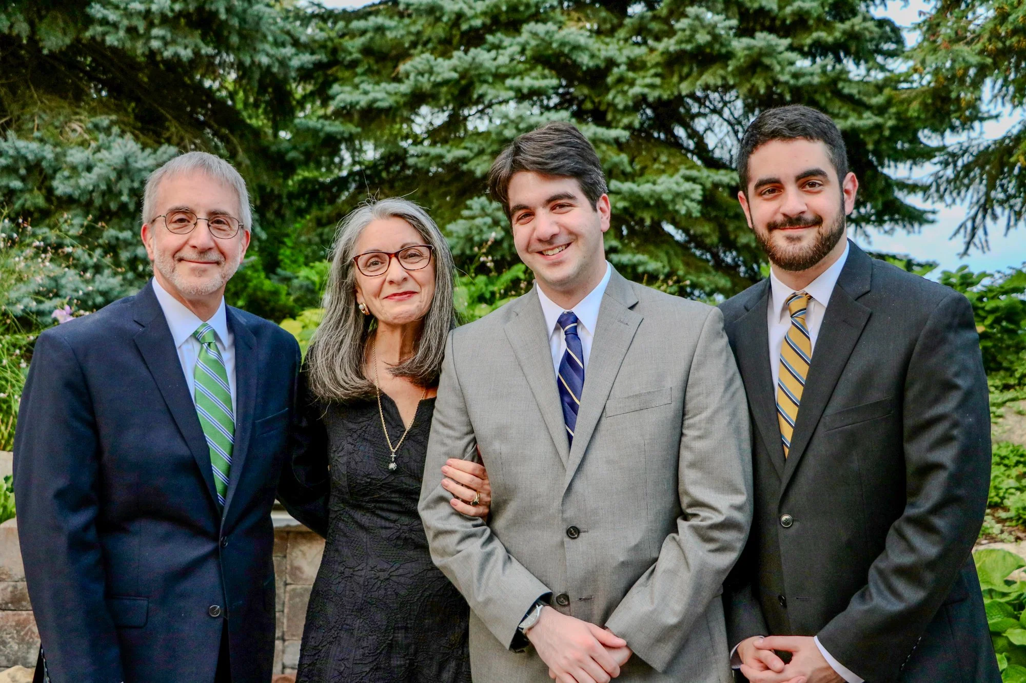 Family portrait of four people, a father, mother, and two adult sons, standing outdoors in front of green trees, dressed in formal attire for a special occasion.