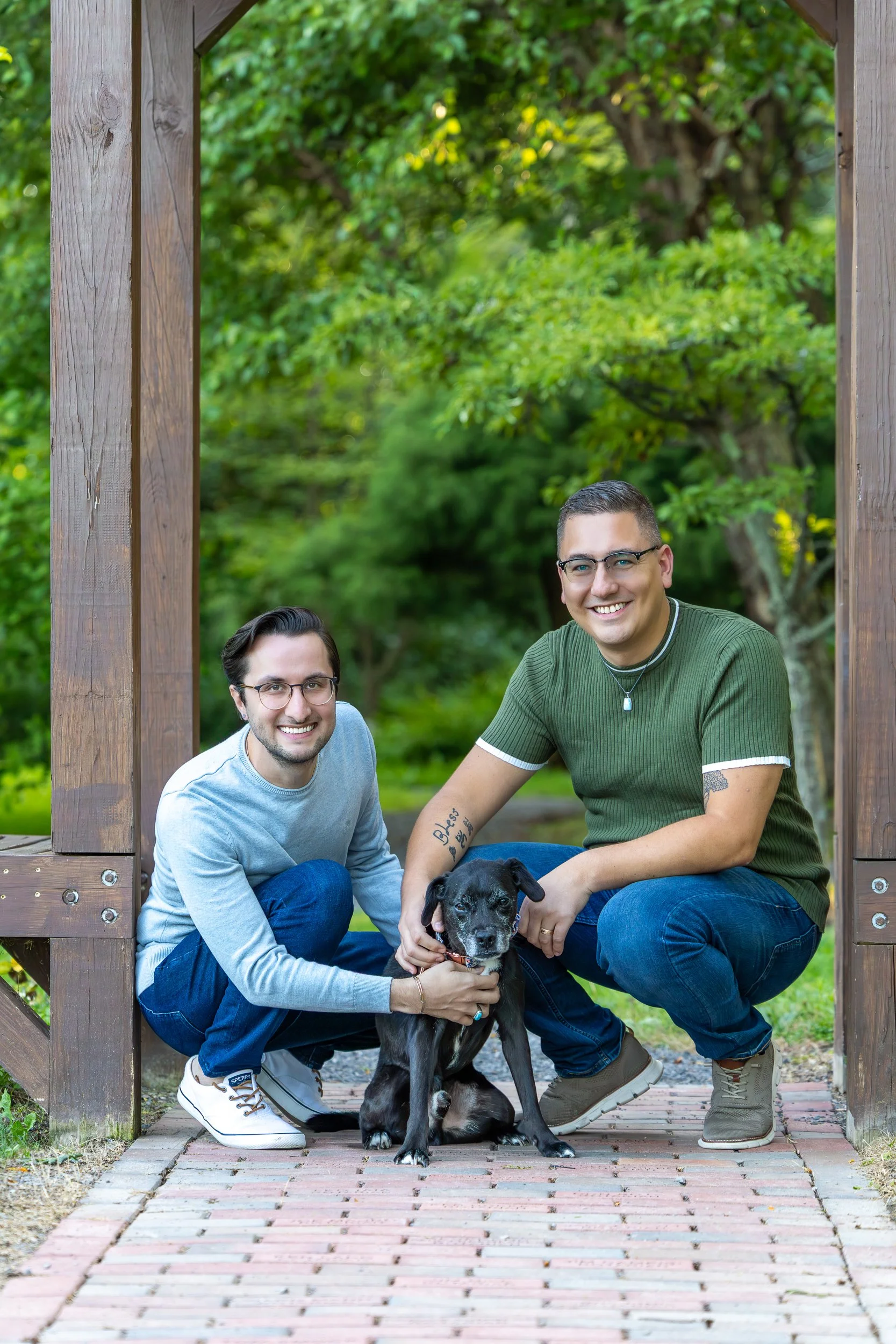 Two smiling men with glasses are squatting on either side of a black dog at the entrance of a wooden archway in a park with green trees in the background.