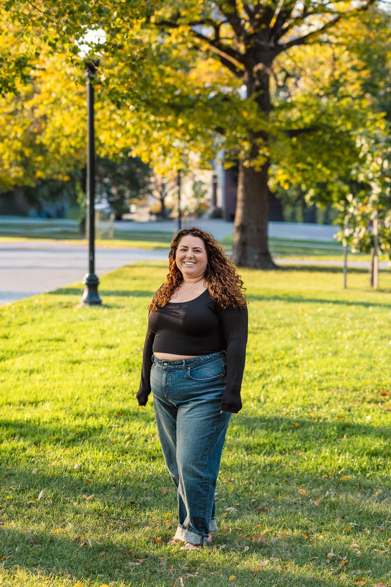 A smiling woman with curly hair walking on a grassy park while wearing a black top and blue jeans, with trees and cars in the background during the daytime.