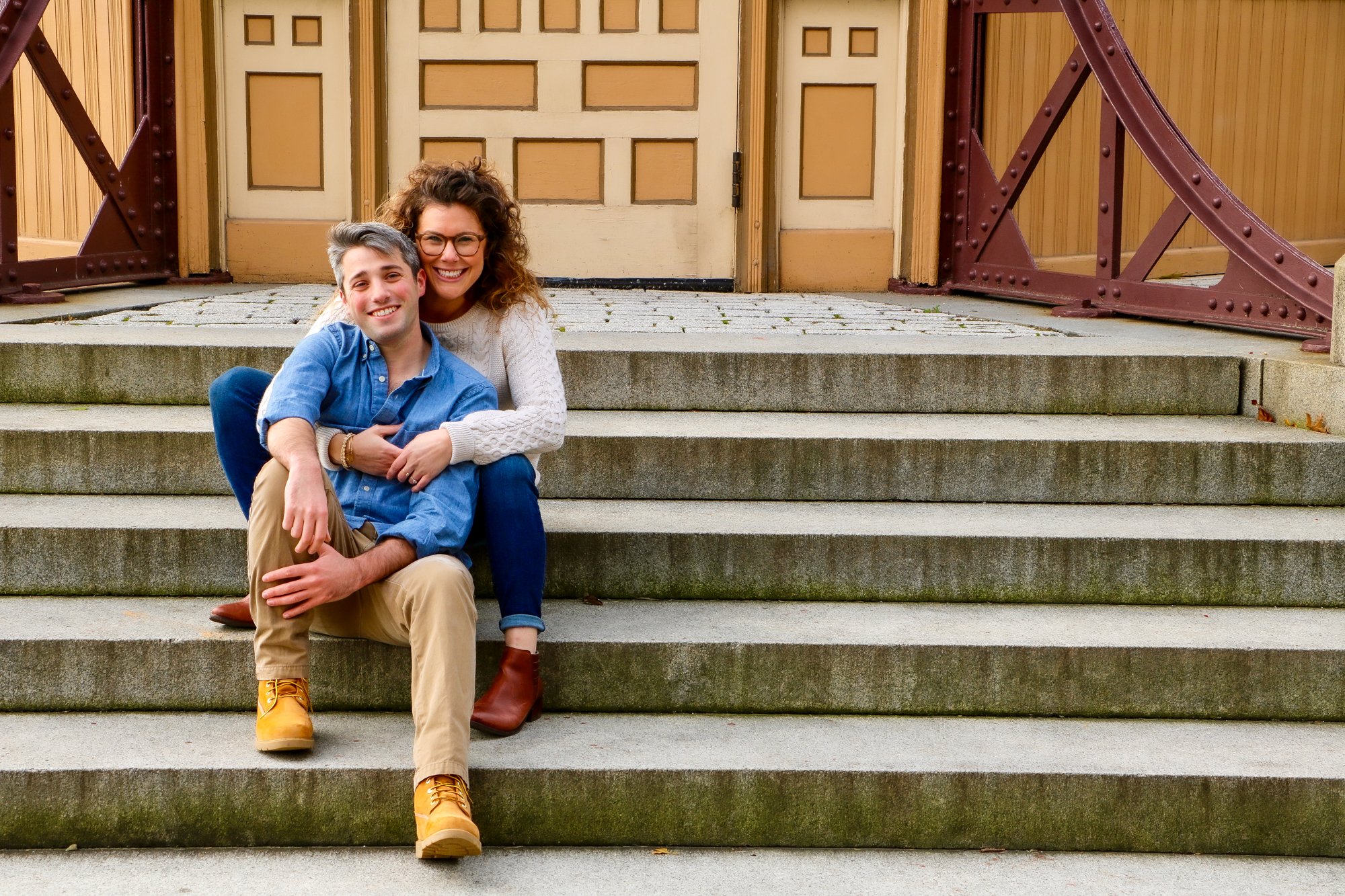 A smiling couple sitting on outdoor stone staircase in front of a beige door, with the woman hugging the man from behind.