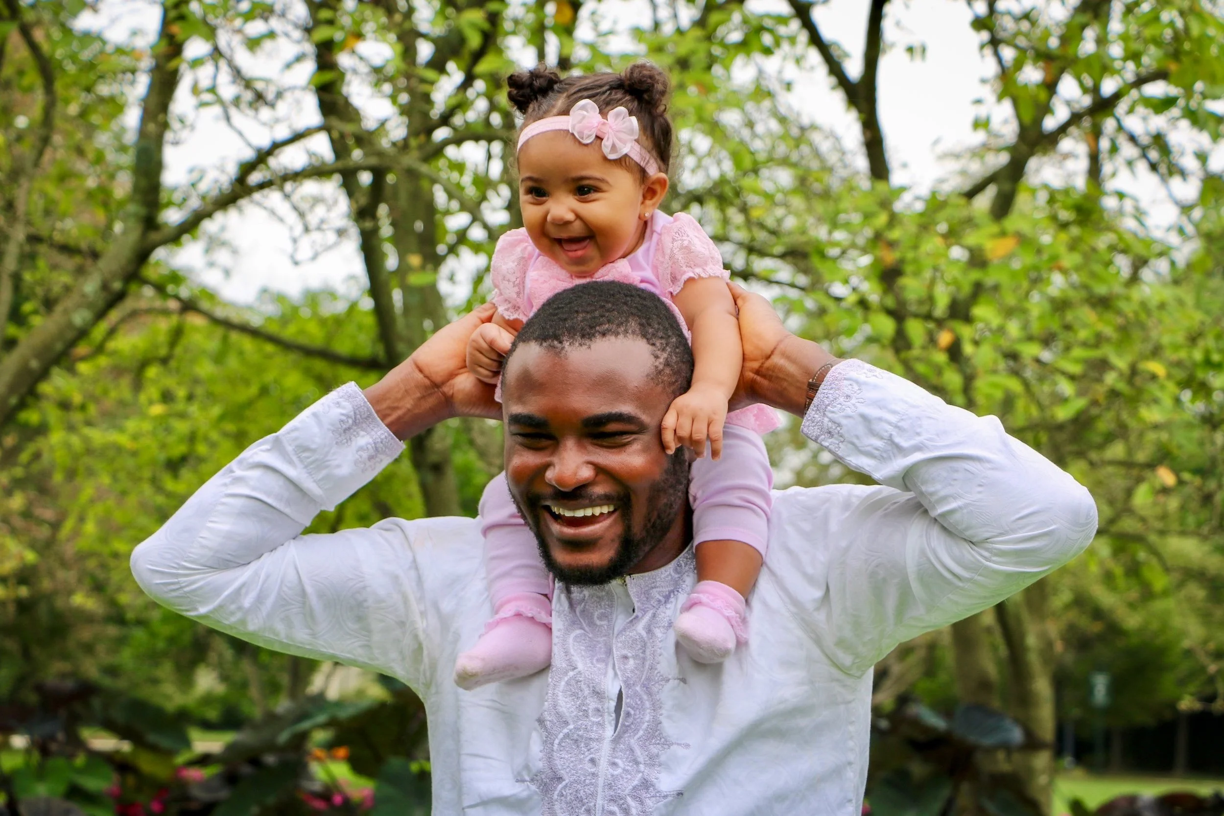 A joyful man carrying a young girl on his shoulders in a park, with green trees and foliage in the background.