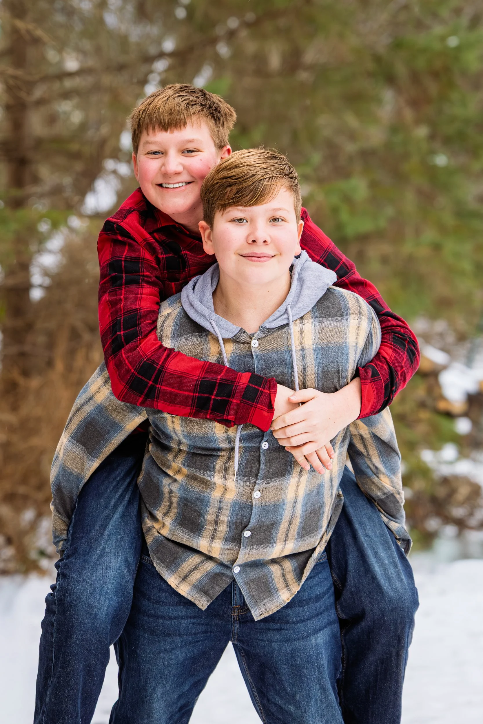 Two boys outdoors in winter, one giving the other a piggyback ride, with trees and snow in the background.