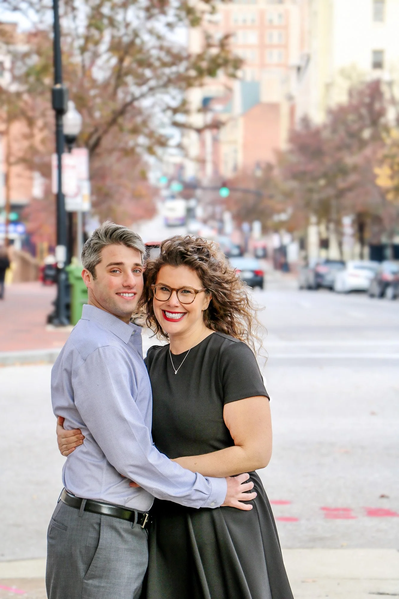 A smiling woman with curly hair and glasses embraces a man on a city street with trees and buildings in the background.