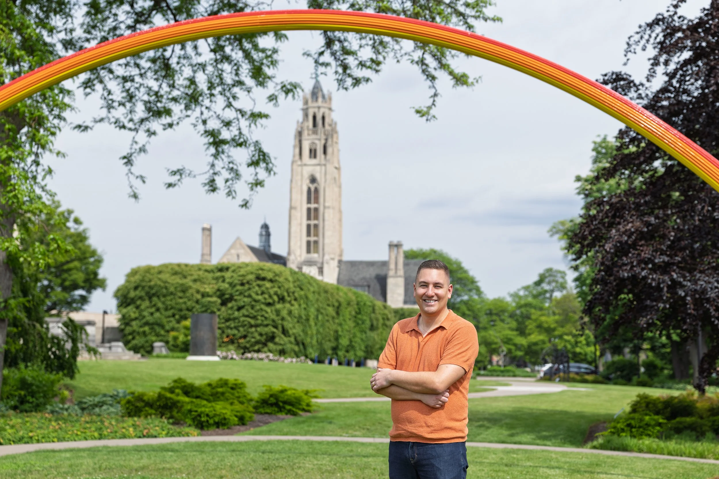 A man smiling with crossed arms standing on a grassy park with thick trees, a church with a tall tower, and a colorful rainbow arch overhead.