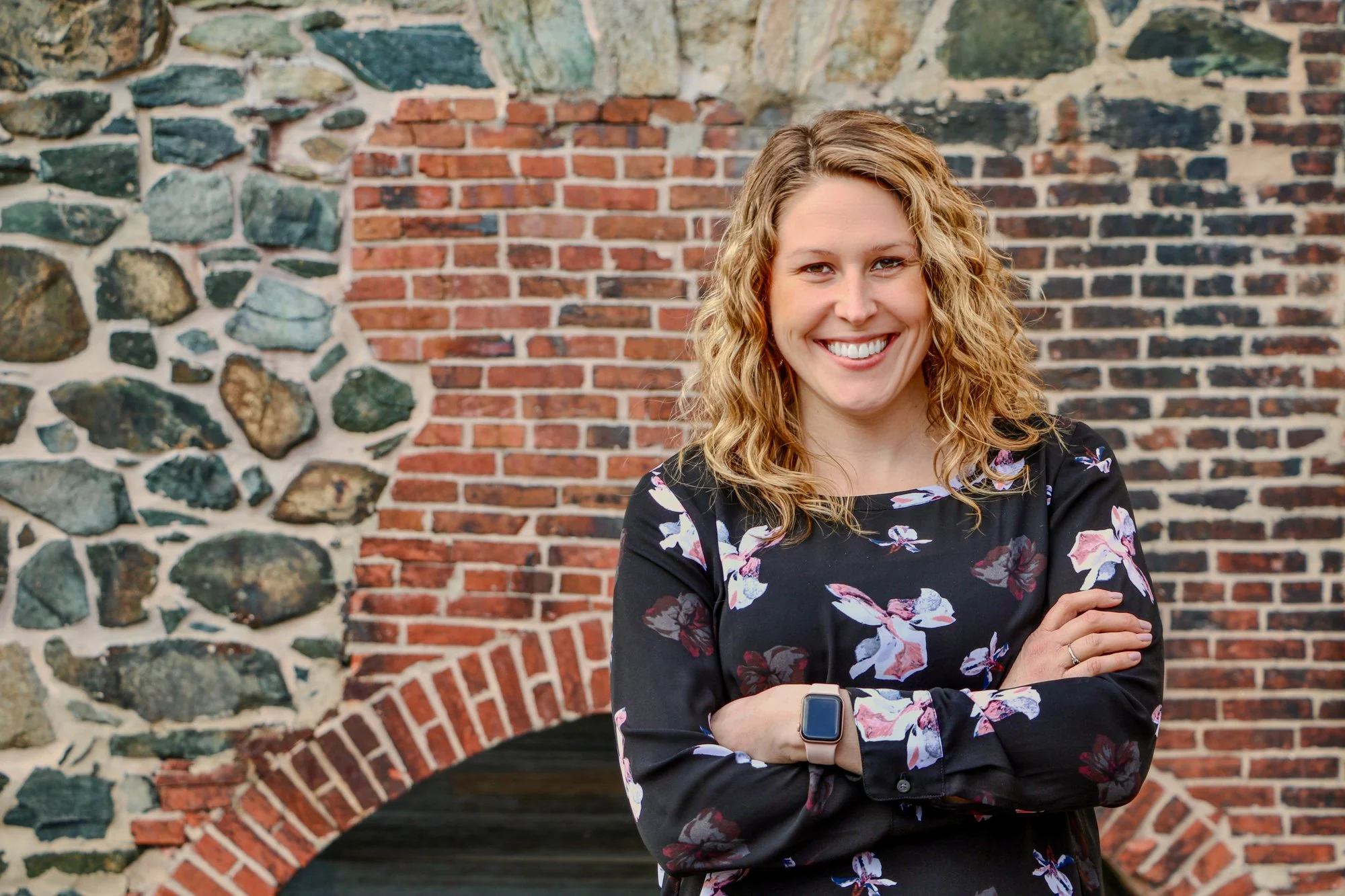 A woman with curly blonde hair, smiling with arms crossed, standing in front of a brick and stone wall.