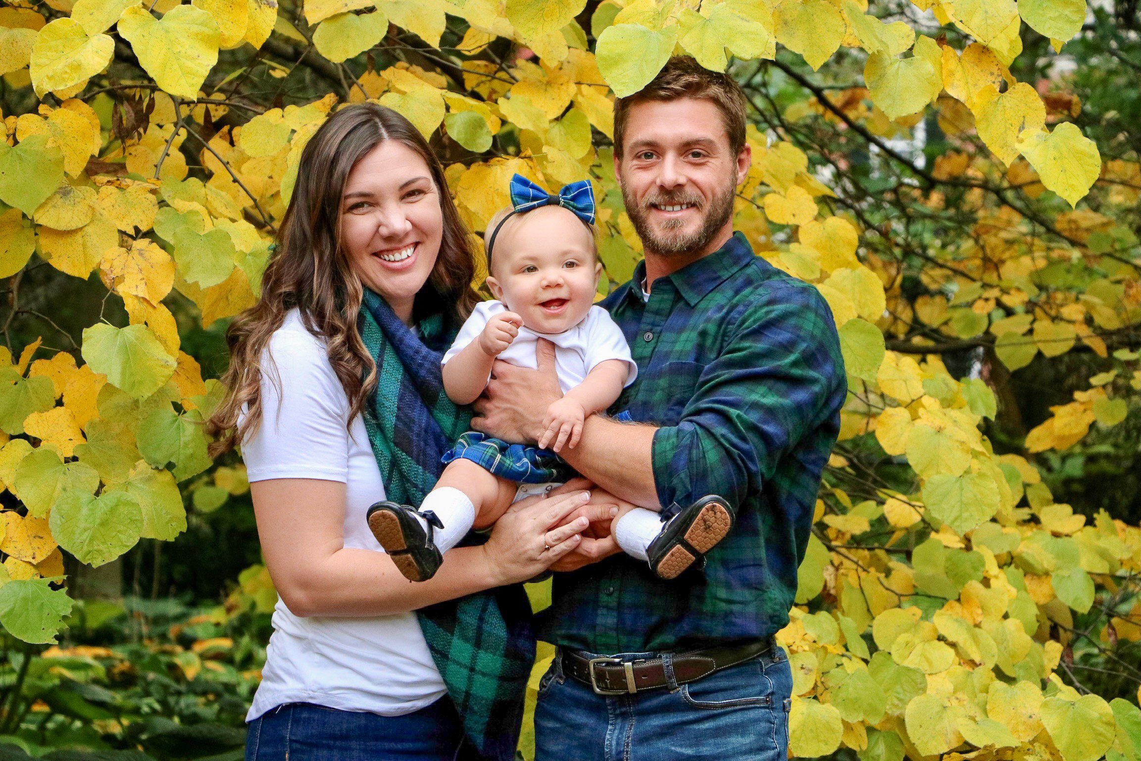 A smiling family of three, a woman, a man, and a baby girl, standing outdoors in front of a tree with yellow leaves, during fall.