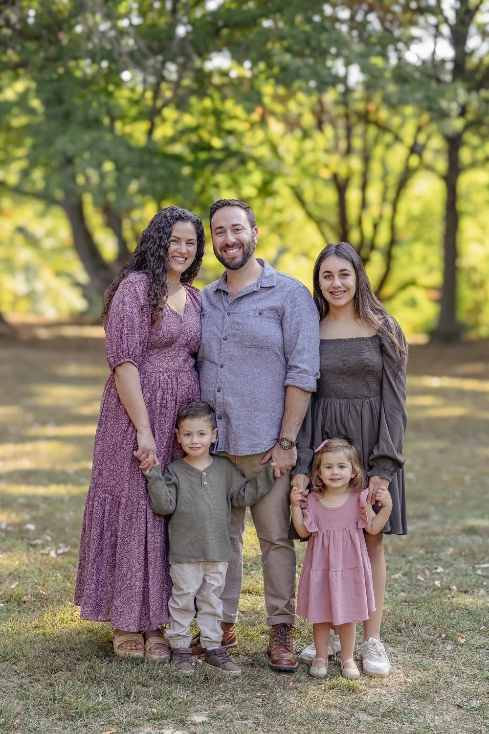 Family of five sitting outdoors in a park during fall, smiling at the camera. The family includes a man with a beard, a woman with curly hair, a teenage girl, a young boy, and a baby girl, with trees and autumn leaves in the background.