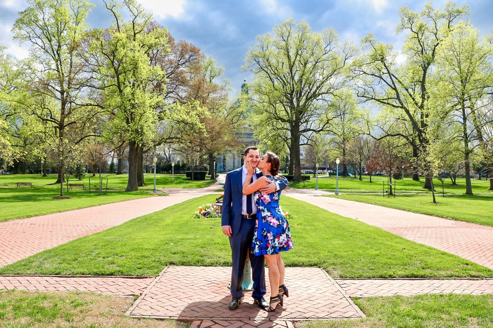 A couple dressed in formal clothes sharing an affectionate moment in a park with green grass and tall trees, with a building and cloudy sky in the background.