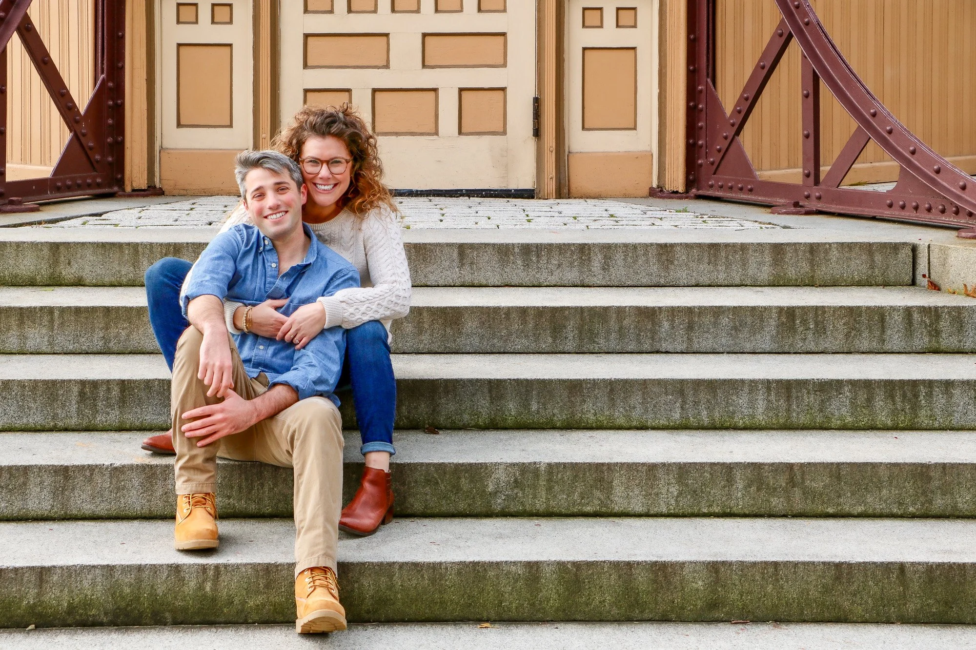 A smiling couple sitting on outdoor stone staircase in front of a beige door, with the woman hugging the man from behind.
