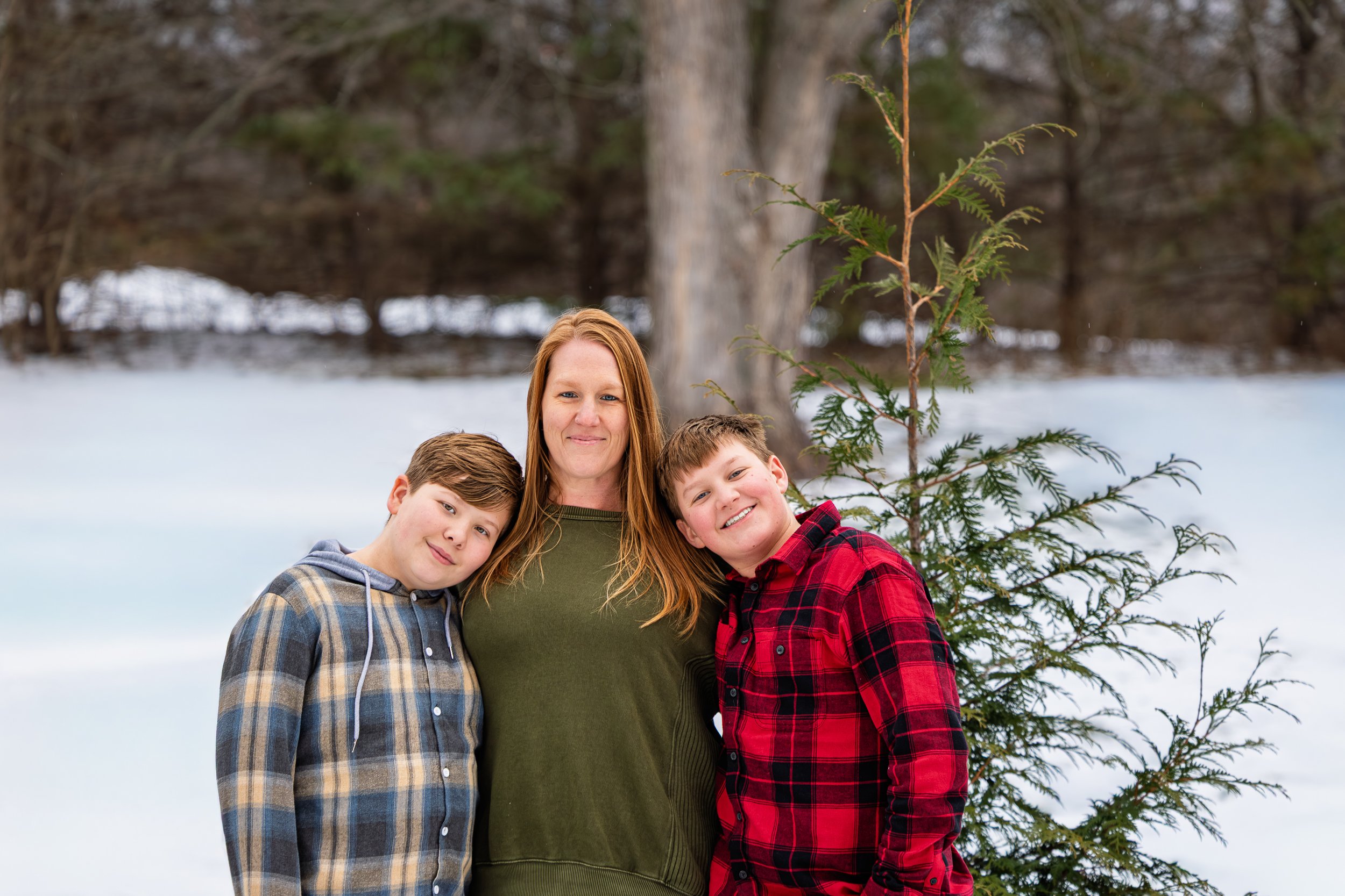 A woman with long auburn hair stands outdoors in a snow-covered area with two young boys, one leaning his head on her shoulder and the other smiling, near a small pine tree, with trees in the background.