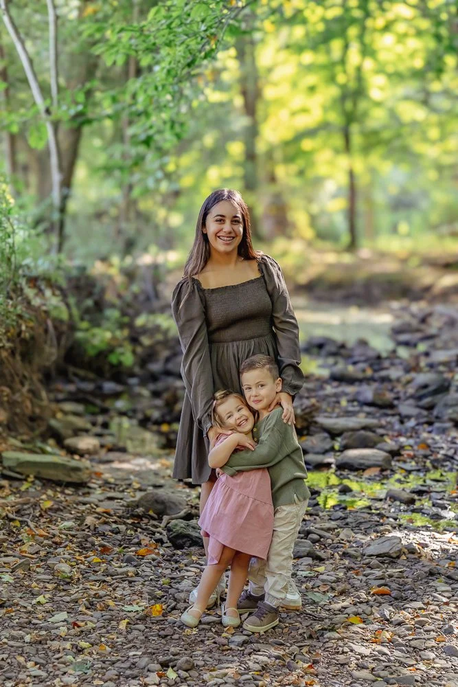 A teenager and two young children standing close together on a rocky path in a forest with green foliage. The woman is smiling and wearing a brown dress, while the children are hugging her, smiling happily.