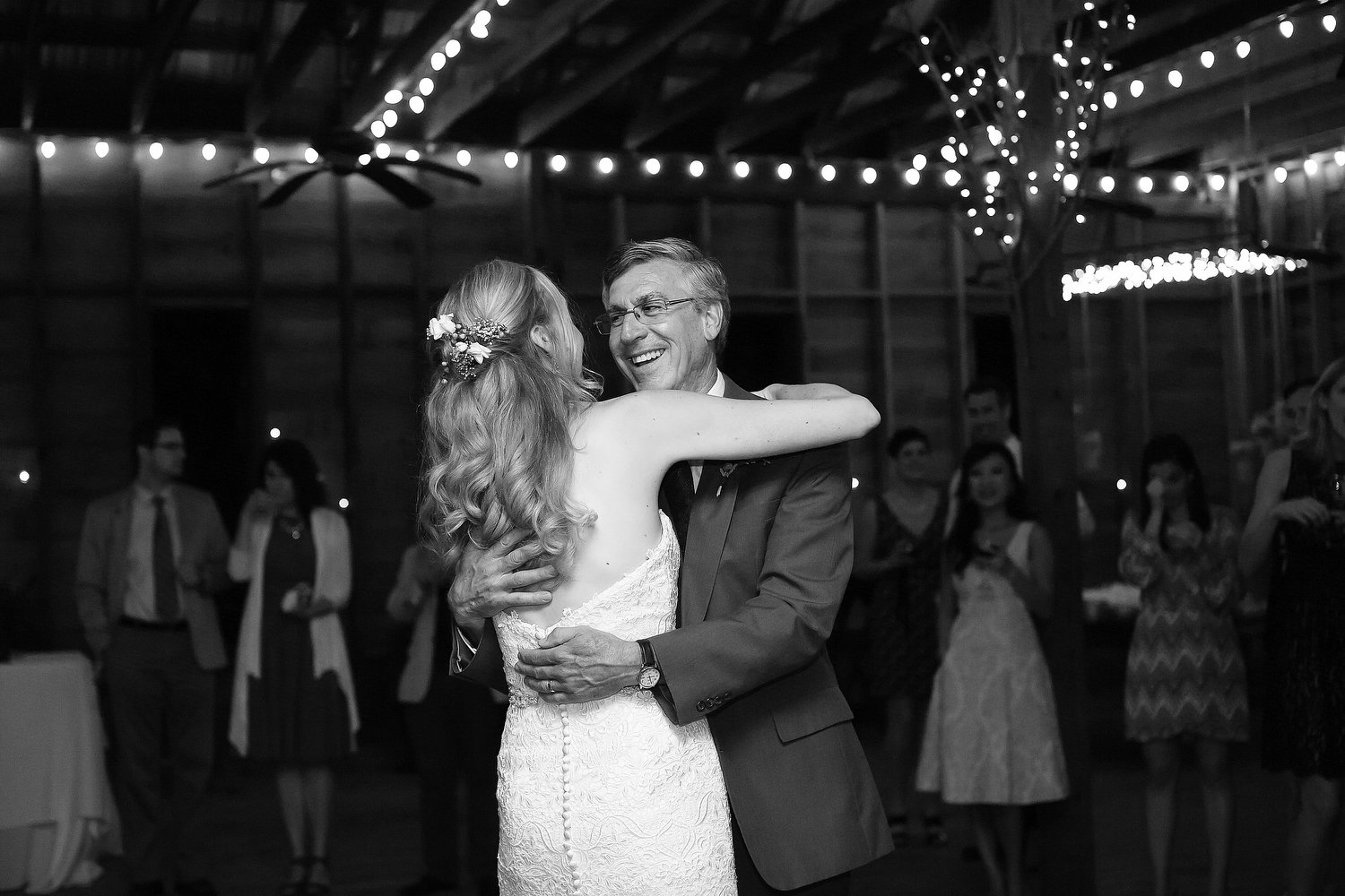 A bride and an older man, possibly her father, sharing a dance at a wedding reception inside a barn decorated with string lights. The bride is in a strapless lace wedding gown with a floral hair accessory, and the man is in a suit with glasses.