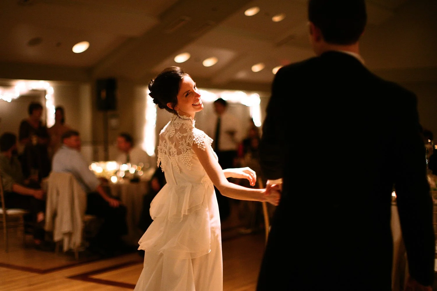 A bride in a vintage white lace dress dancing with a groom in a black suit at a dimly-lit wedding reception, with friends and family seated at tables in the background.