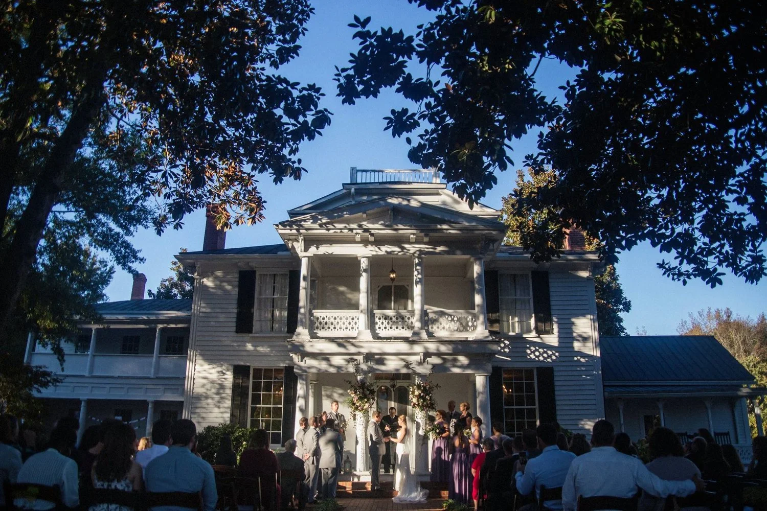 An outdoor wedding ceremony taking place in front of a large, historic, white wooden house with a wraparound porch and multiple windows. People are seated on chairs, and the bride and groom are standing at the altar with officiant, surrounded by frie