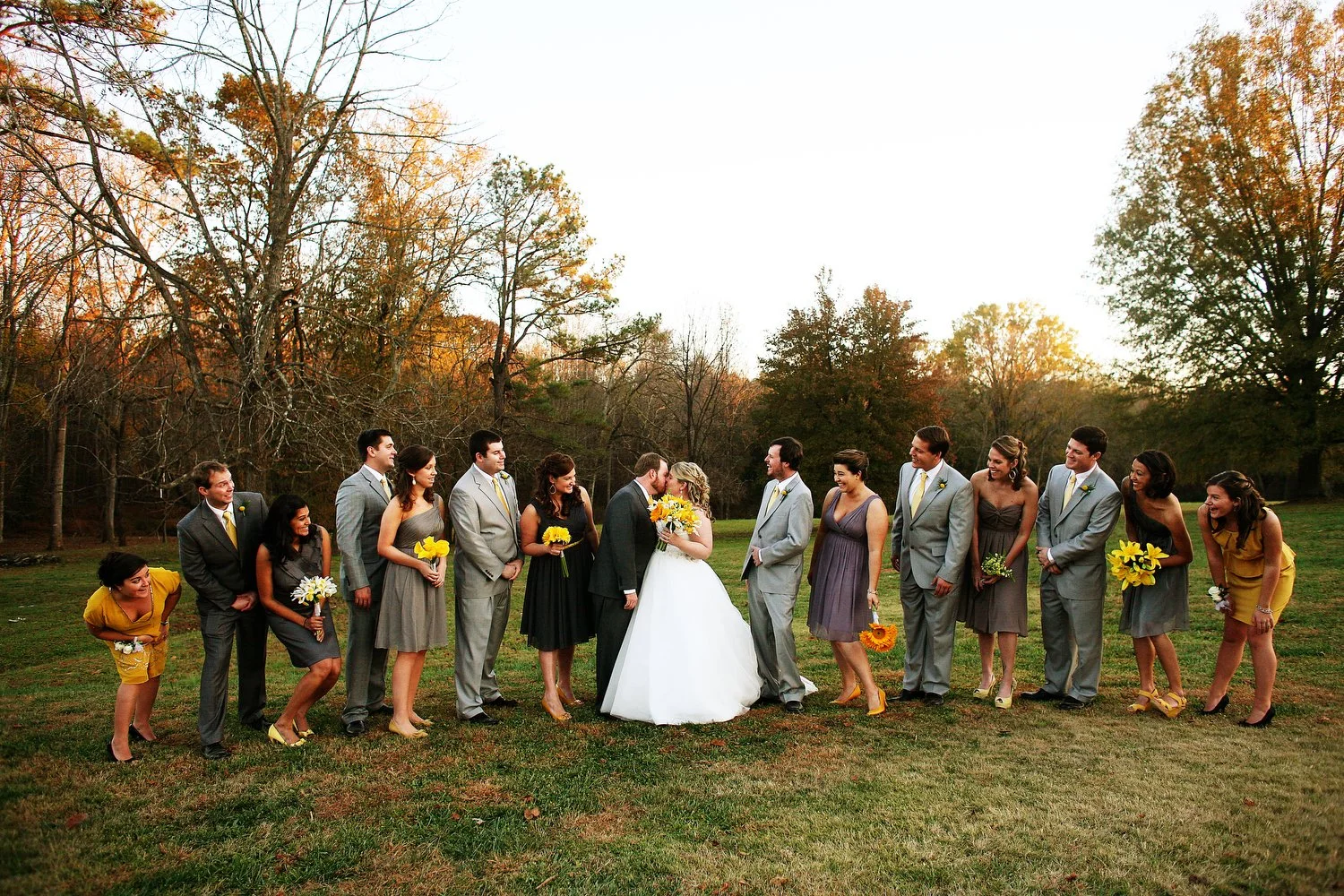 A wedding party standing outdoors on grass with fall trees in the background, celebrating as the bride and groom kiss in the center, surrounded by their family and friends in formal attire, some holding bouquets.