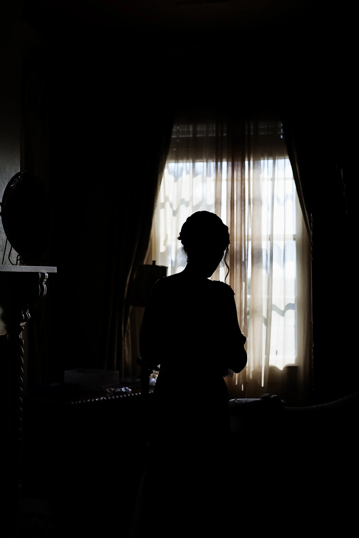 Silhouette of a woman standing near a window with curtains, backlit by daylight.