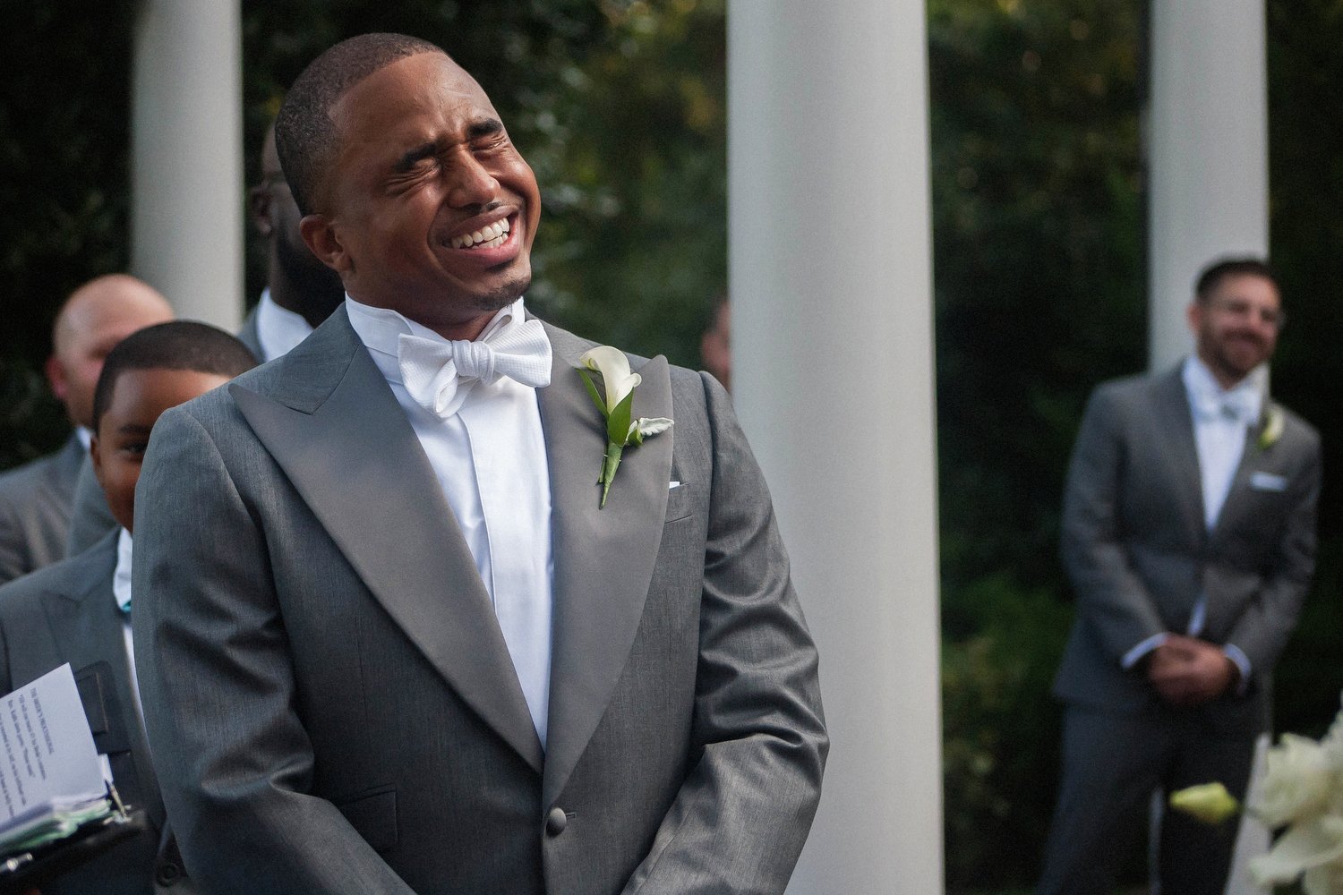 Groom in a gray tuxedo with white shirt and bow tie, smiles emotionally with eyes closed, during a ceremony. He has a white calla lily boutonniere on his lapel. Behind him, others in similar tuxedos are visible, standing outside near white columns.  
