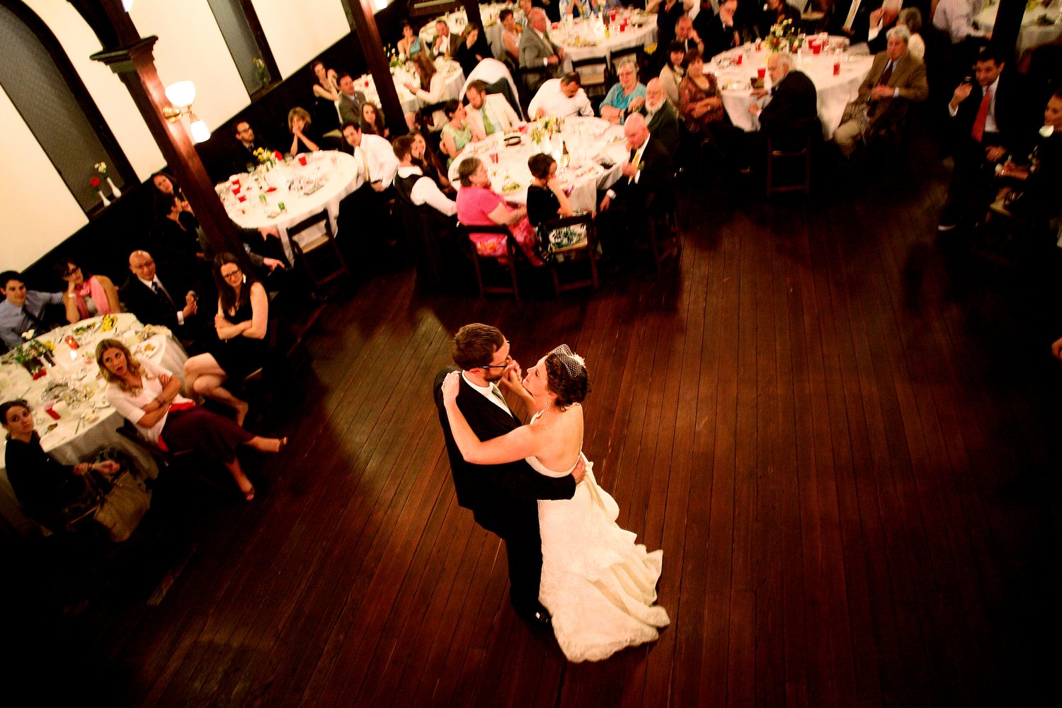 Aerial photo of a bride and groom dancing at their wedding reception, surrounded by seated guests at decorated tables in a formal setting with wooden floors.