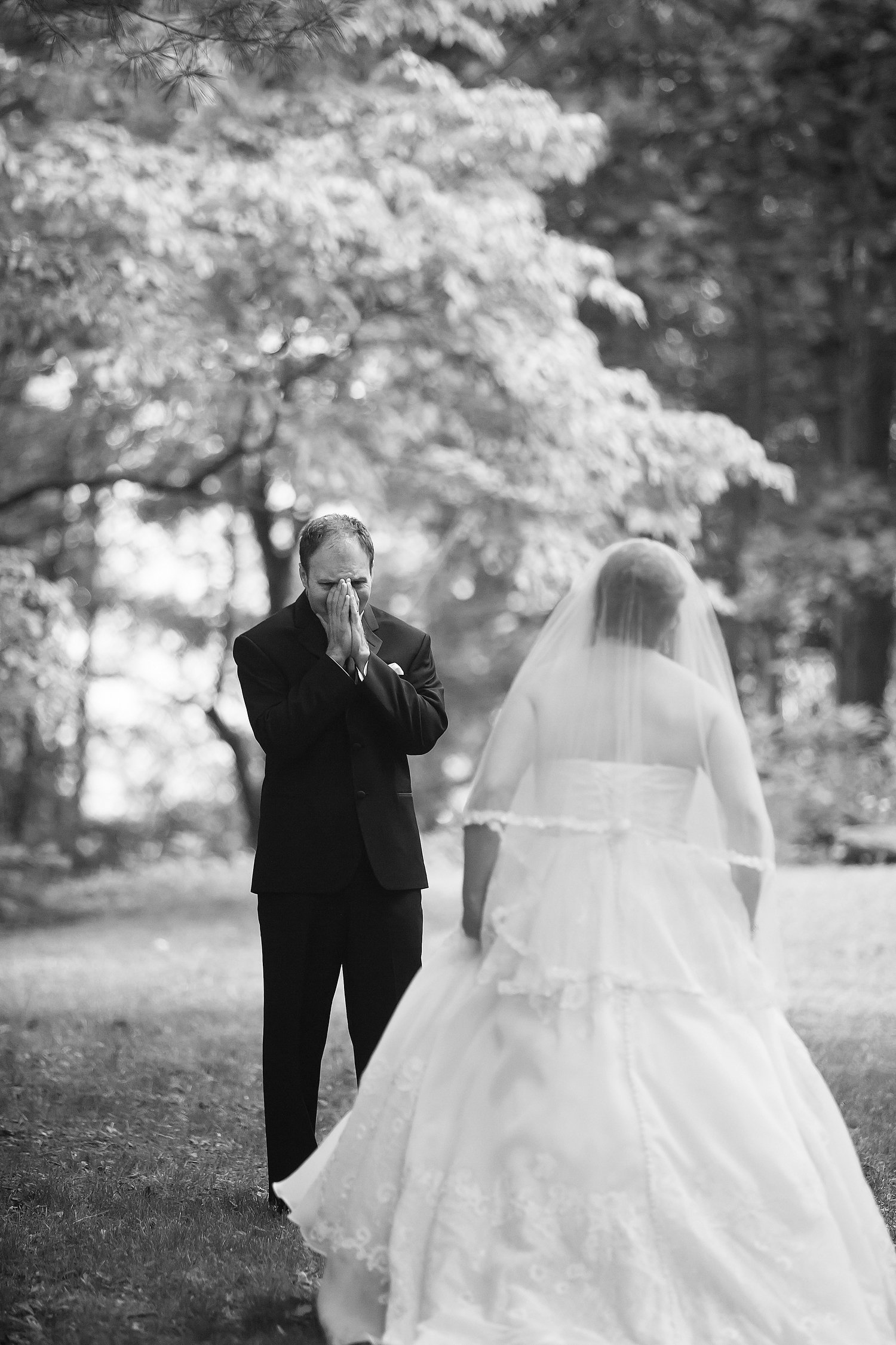 A groom in a tuxedo with his hands covering his face, appearing emotional, facing a bride in a wedding dress with a veil outdoors in a wooded area.