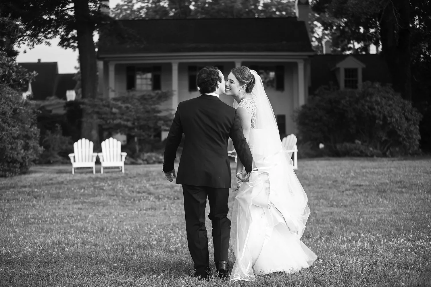 Black and white photo of a bride and groom walking hand in hand on a lawn, smiling at each other, with a house and trees in the background. She has a white gown and a long white veil. He has a black tuxedo.