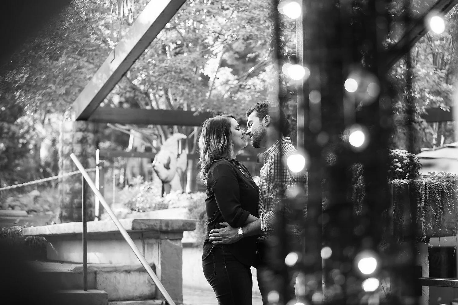 Black and white photo taken from around the corner of a wooden outdoor structure. A couple is seen embracing and kissing on the other side of the structure. Trees are in the far distance. 
