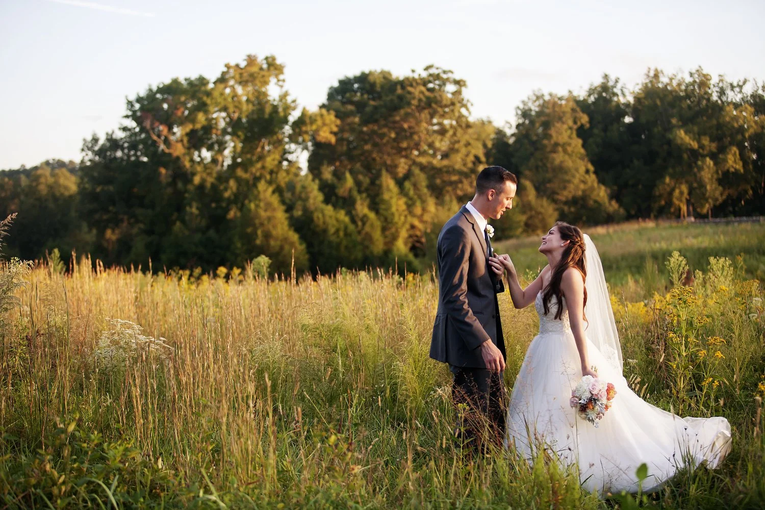 A bride and groom standing in a field of tall grass, with trees in the background, during sunset. The bride is wearing a white wedding dress and holding a bouquet, and the groom is in a gray suit. They are looking at each other happily.