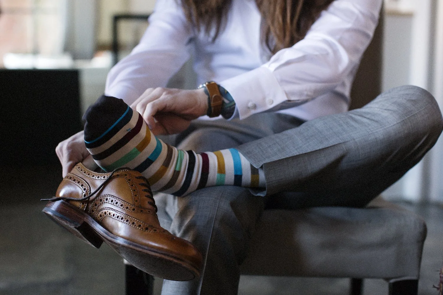 A man adjusts his multicolored striped sock while sitting on a gray armchair, wearing brown leather brogue shoes, a white dress shirt, gray plaid pants, and a wristwatch. His face is cut off at the top of the photo.