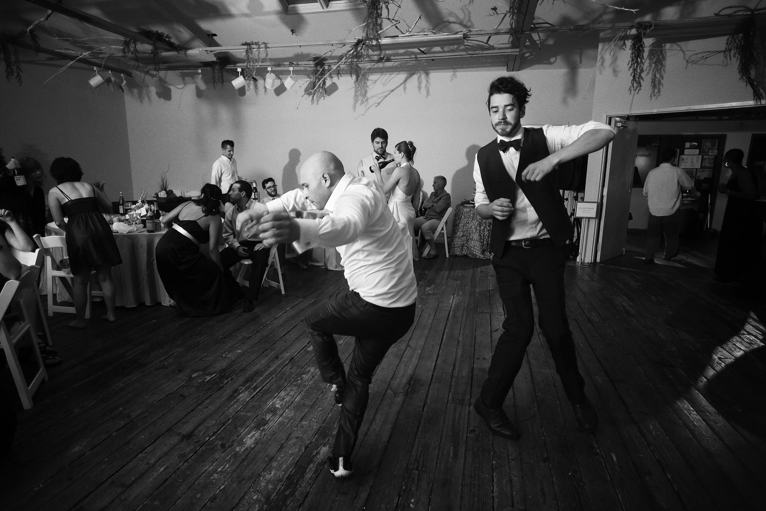Black and white photo of a dance floor. The room is dimly lit. People are seated at tables in the background. Two men twist and bend their arms on the dance floor. One is bald and wearing a white shirt and black pants. The other has dark hair and is 